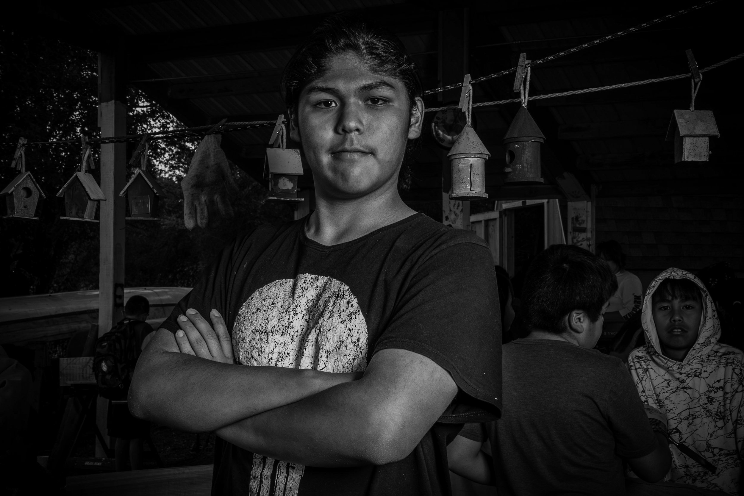 A young man standing with arms crossed, wearing a T-shirt with a skull design, in front of a group of children and hanging birdhouses. The setting appears to be outdoors with a wooden structure overhead.