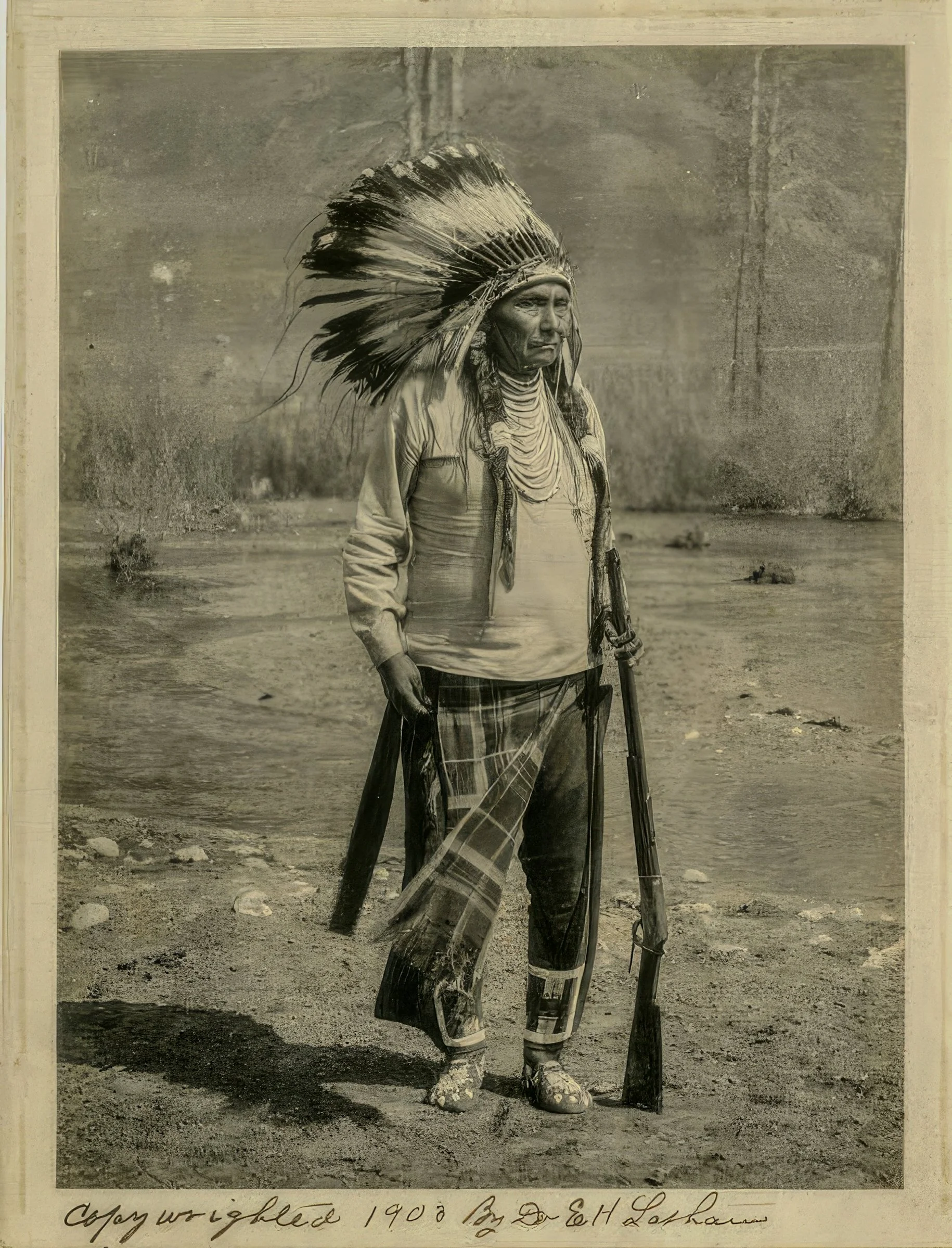 A black and white photograph of a Native American man wearing a large feathered headdress and traditional clothing, holding a staff or walking stick, standing outdoors on dirt ground with a plain background.