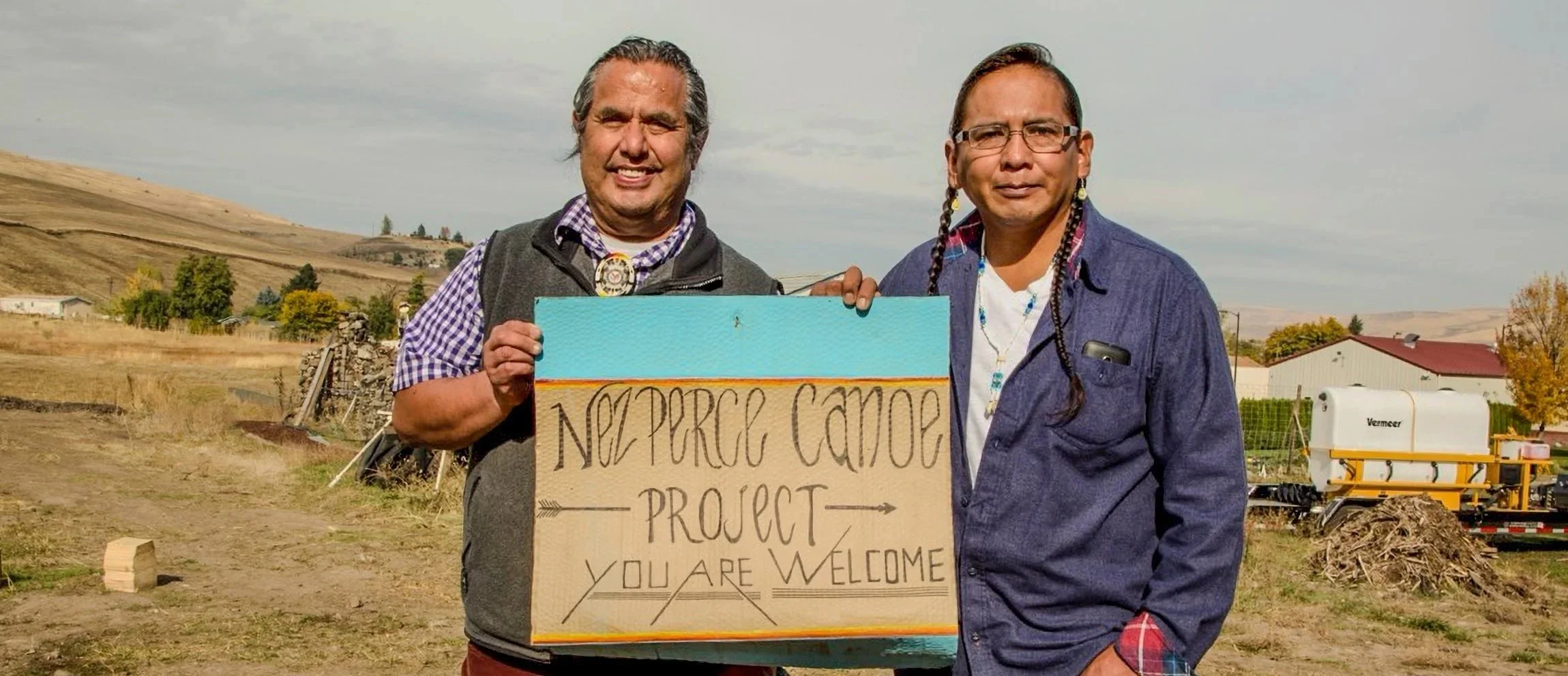 Two men standing outdoors, holding a sign that reads "Neppeace Camp Project - You Are Welcome". One man is smiling and wearing a checkered shirt with a vest, the other man is serious and wearing glasses and a denim jacket. The background shows a rural landscape with hills, trees, a few buildings, and farm equipment.