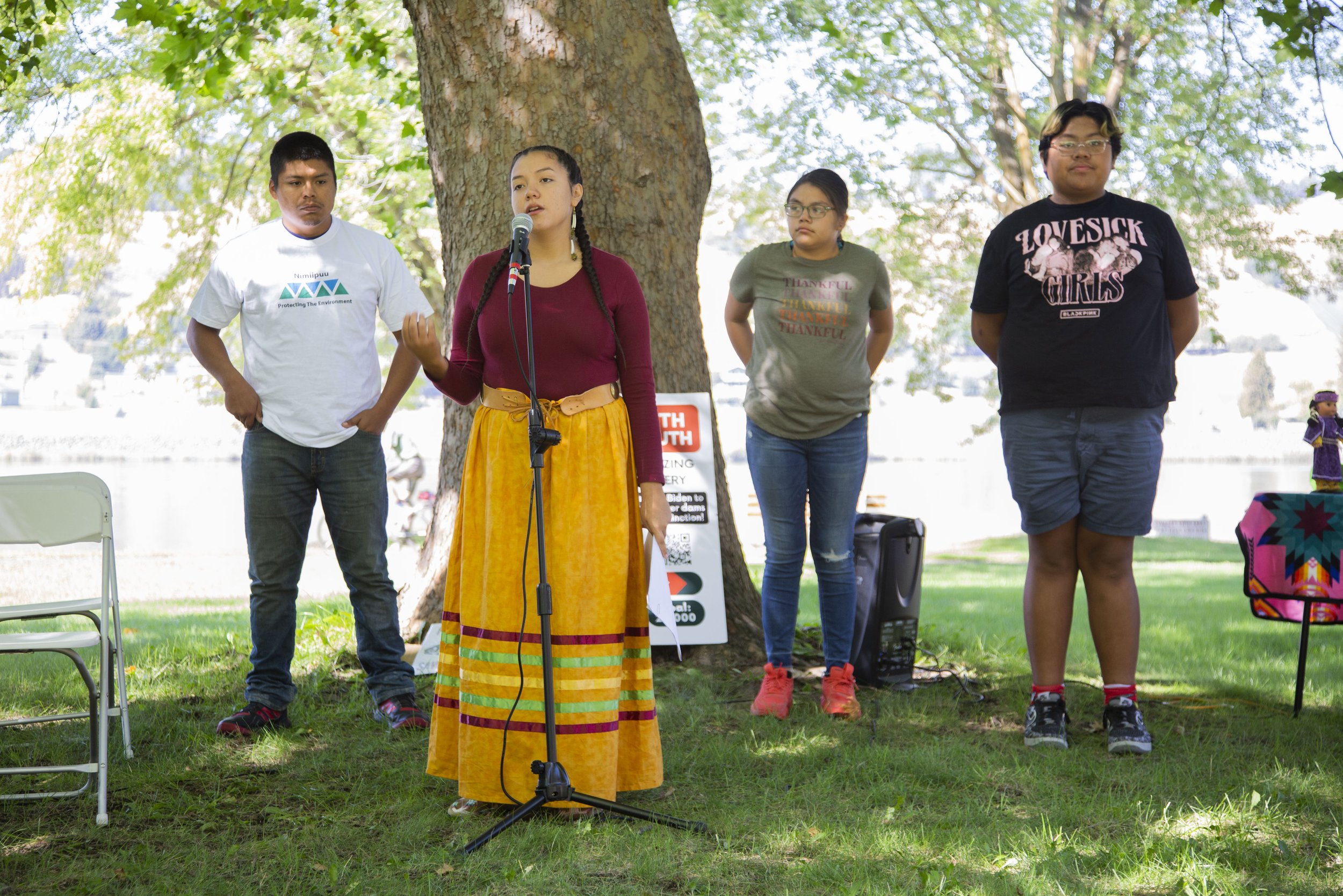 A woman with braided hair wearing a maroon top and a long yellow skirt with colorful stripes is speaking into a microphone outdoors, standing in front of a large tree. Four people stand behind her, including a man in a white shirt, two women in t-shi