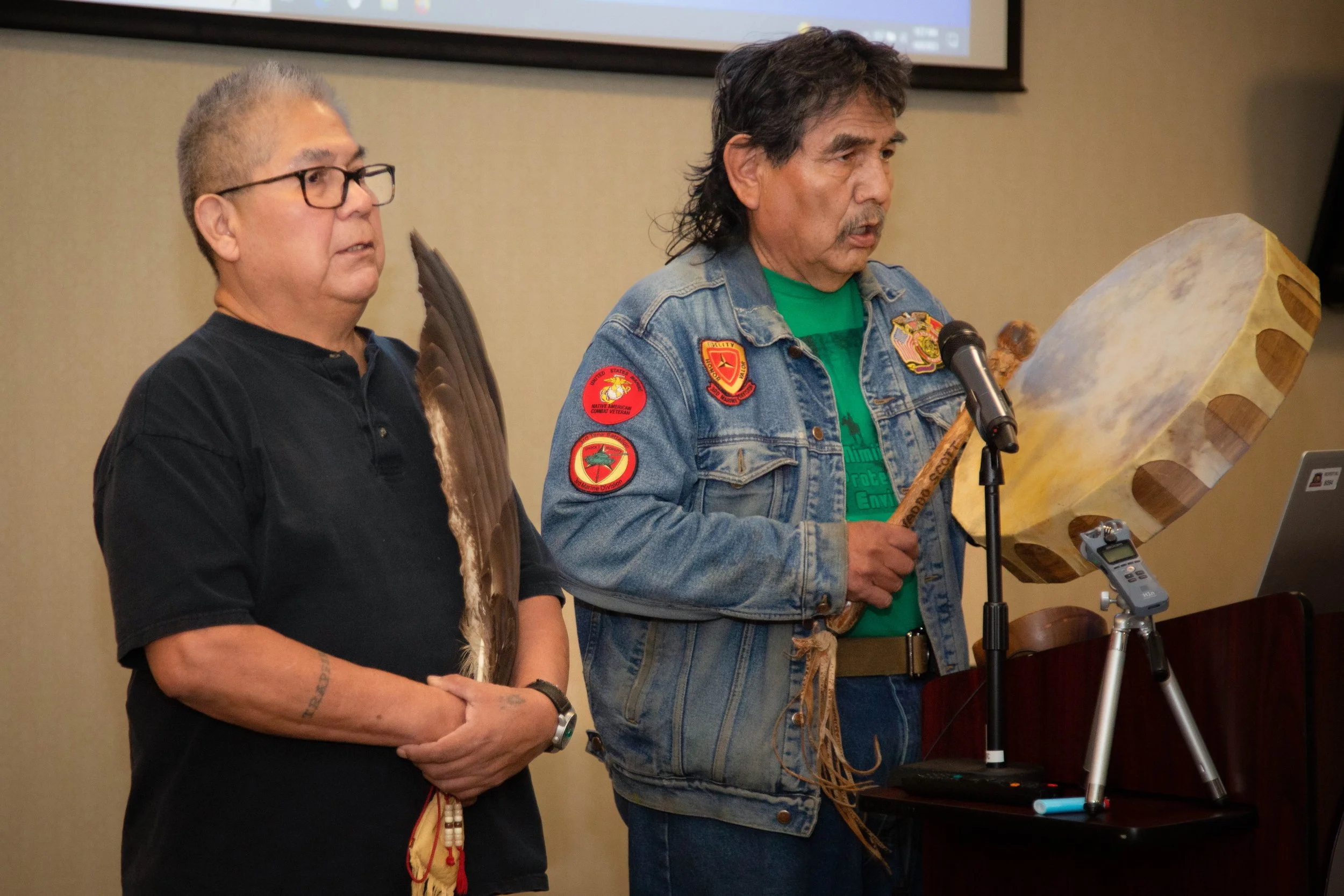 Two men stand in front of a screen, in a room with beige walls, participating in a presentation or ceremony. One man is wearing a black shirt and glasses, holding a wooden staff; the other man, in a denim jacket with patches, is speaking into a microphone while holding a carved wooden staff.
