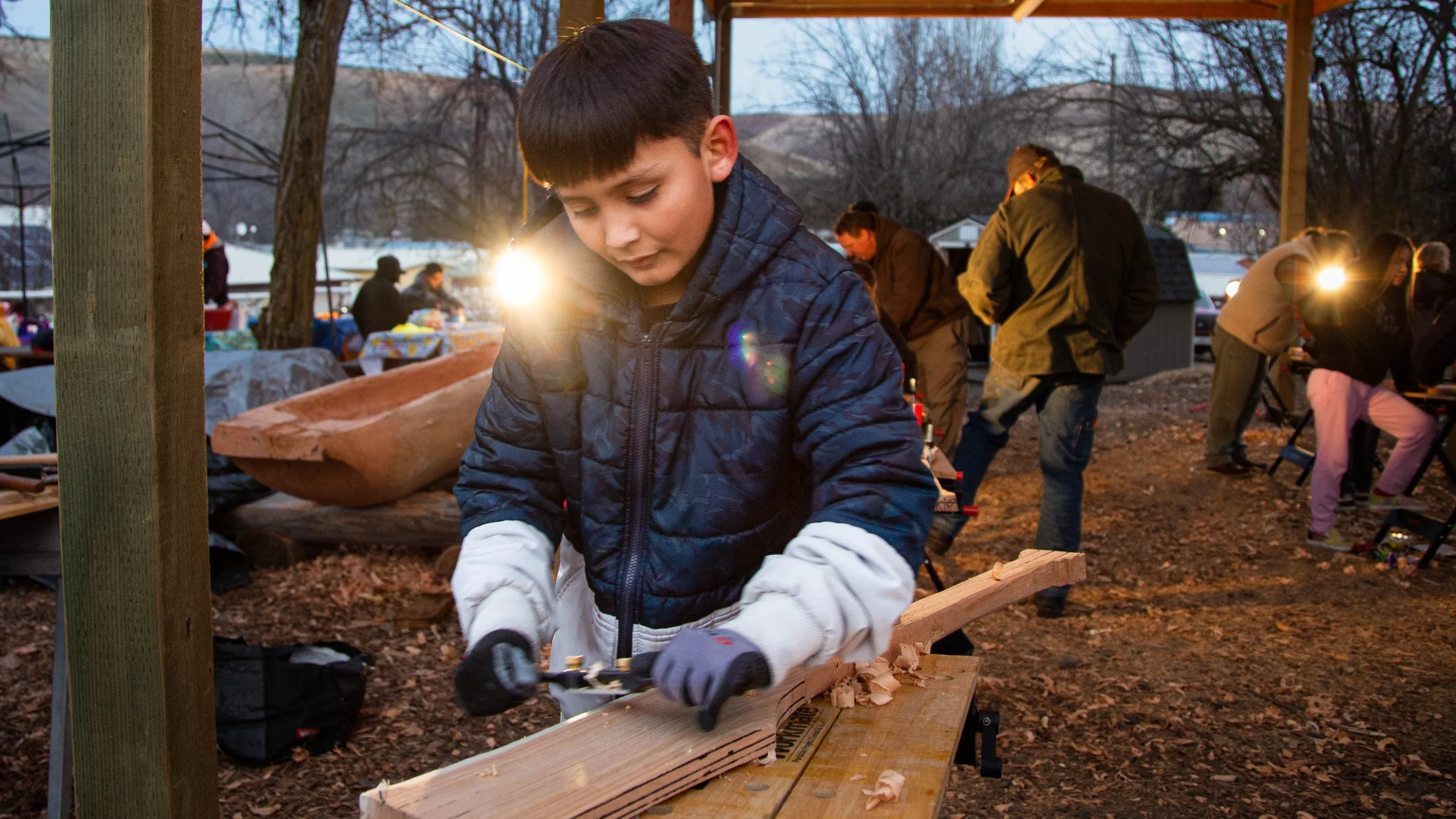 A young boy in a blue jacket using a hand saw to cut a piece of wood at an outdoor gathering during the evening. In the background, people are gathered around tables, some looking at their phones, with trees and parked cars visible.