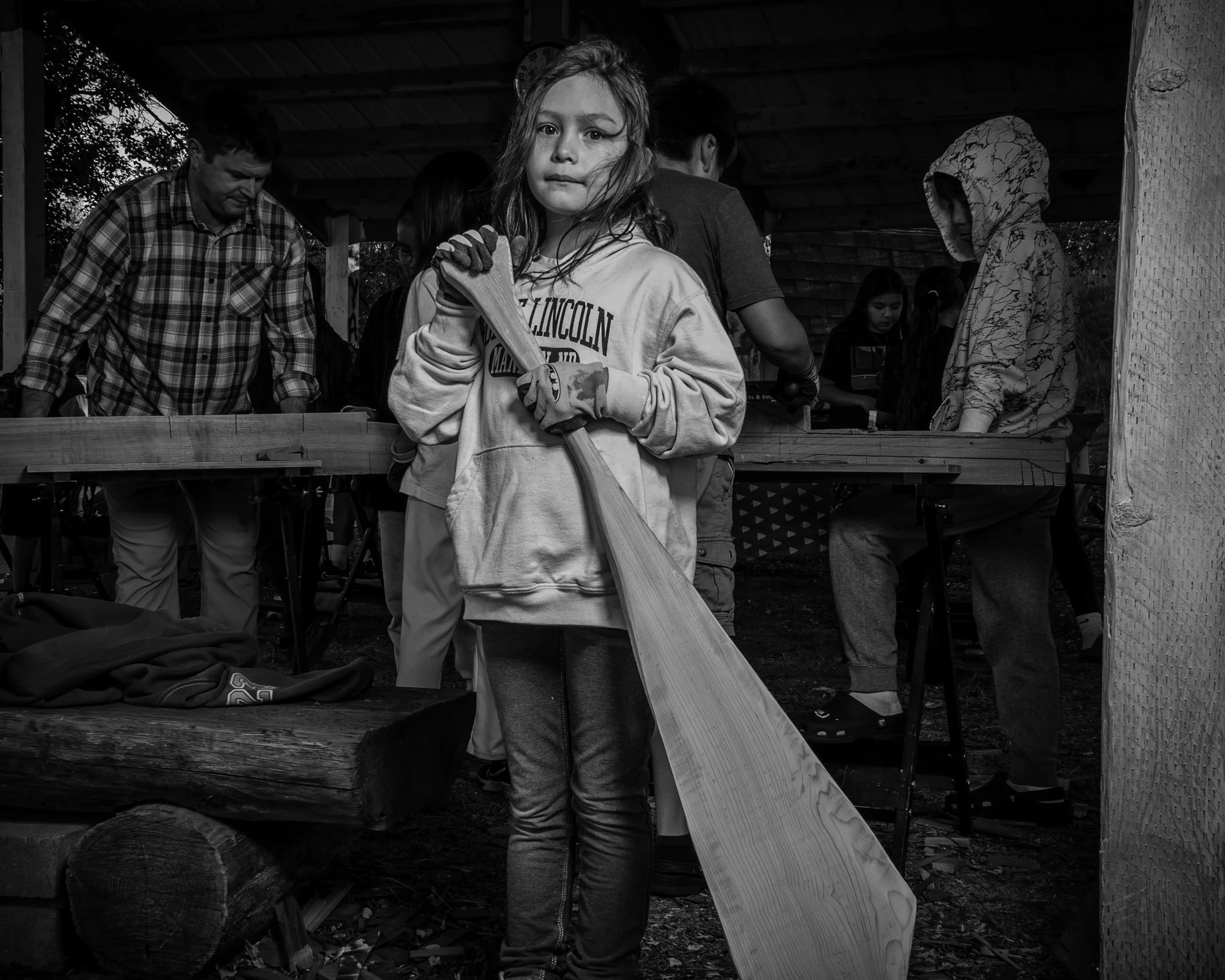 A young girl with wet hair holding a large hammer, standing in front of a group of people at an outdoor event under a wooden shelter. The scene is in black and white.