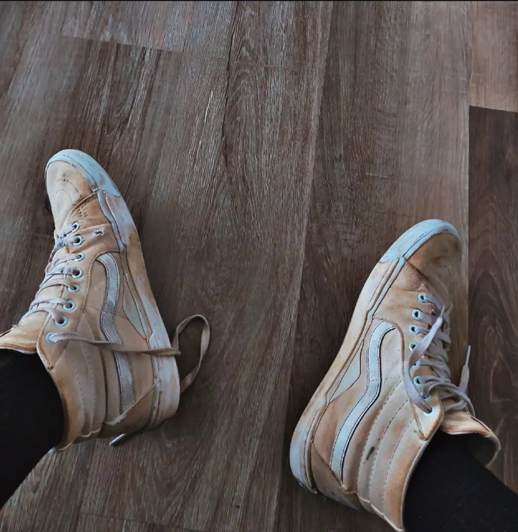 A person wearing beige high-top sneakers with white soles, sitting on a brown wooden floor.