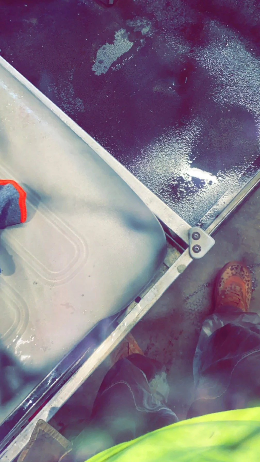 Close-up view of a black, frosted glass surface with water spots, viewed from above, with a corner of a metallic table or work surface and a section of a person's leg and orange work boots visible.