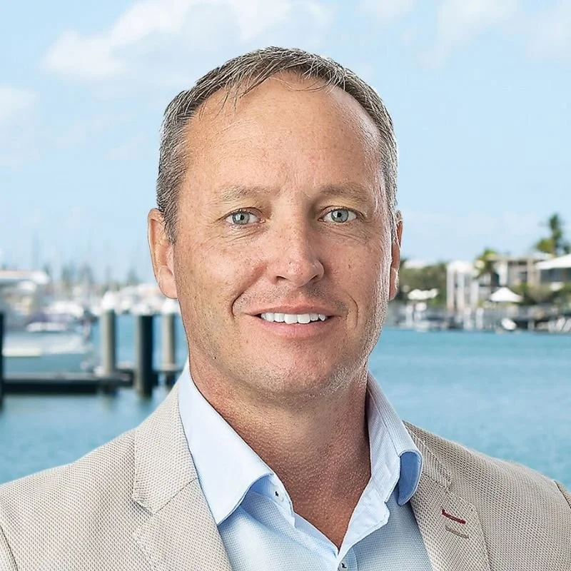 Smiling man in a beige suit jacket and light blue shirt standing outdoors near a marina with boats and blue sky in the background.