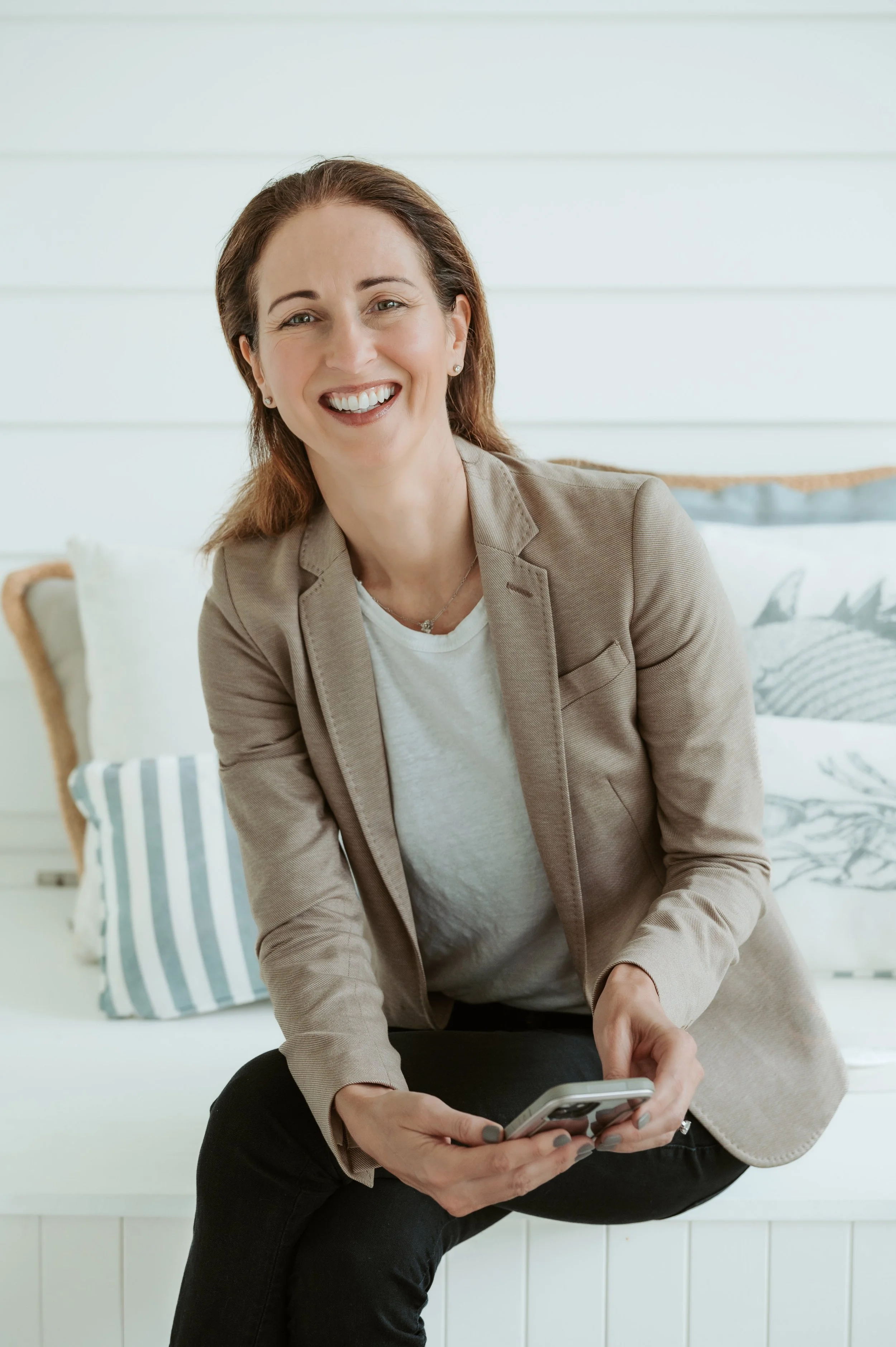 Smiling woman in a beige blazer and white shirt sitting on a white bench with pillows, holding a smartphone.
