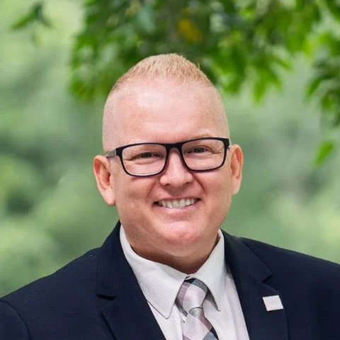 Portrait of a smiling man with glasses and short blonde hair, wearing a dark blazer, light shirt, and patterned tie, outdoors with green foliage in the background.