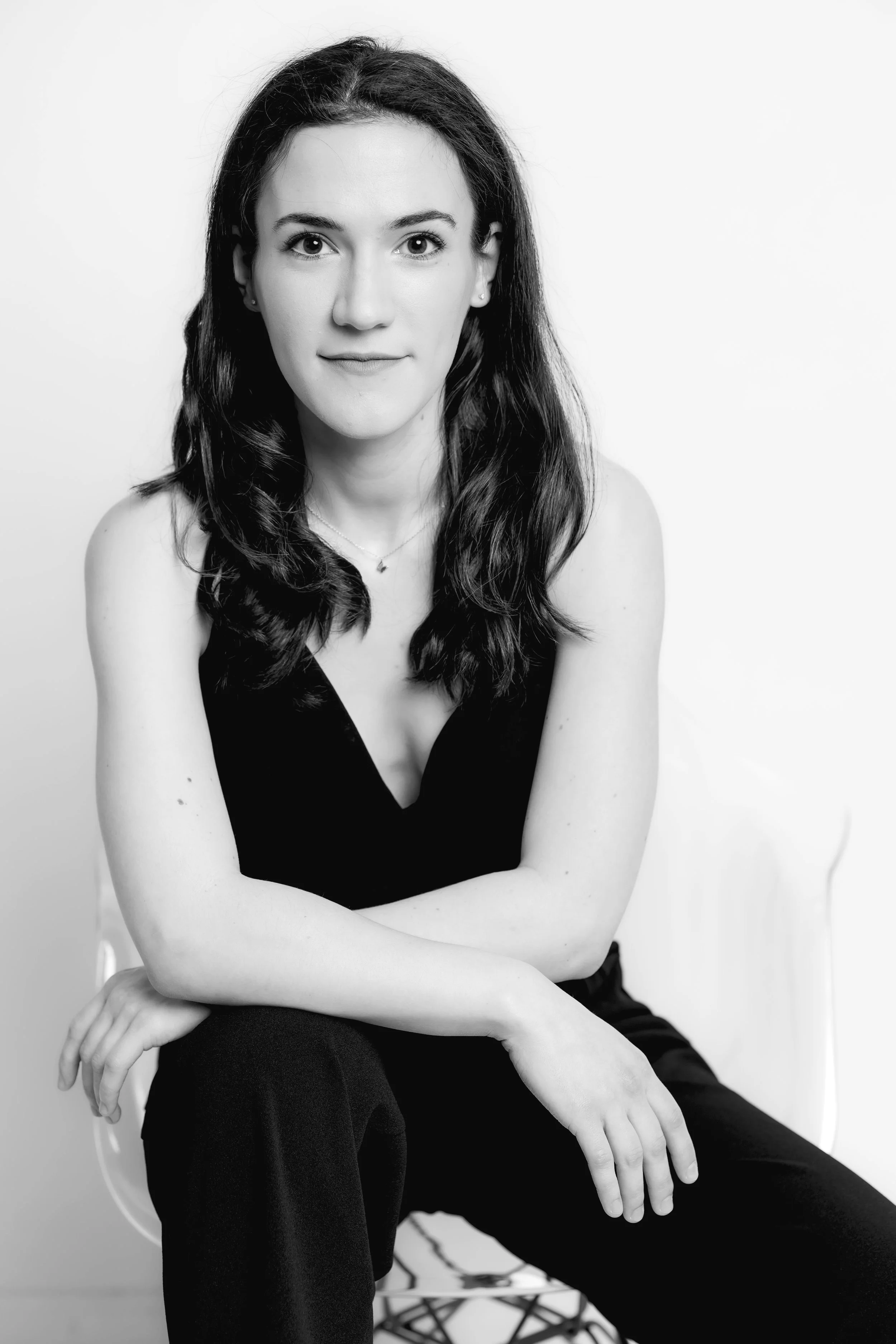 A black-and-white portrait of a woman with long, wavy dark hair, wearing a sleeveless black top, sitting on a white chair against a plain background.