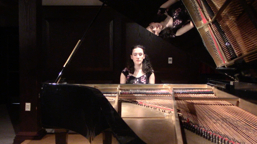 A woman sitting at a grand piano in a dark room, with the piano's inside and strings visible, and her reflection showing on the opened lid.