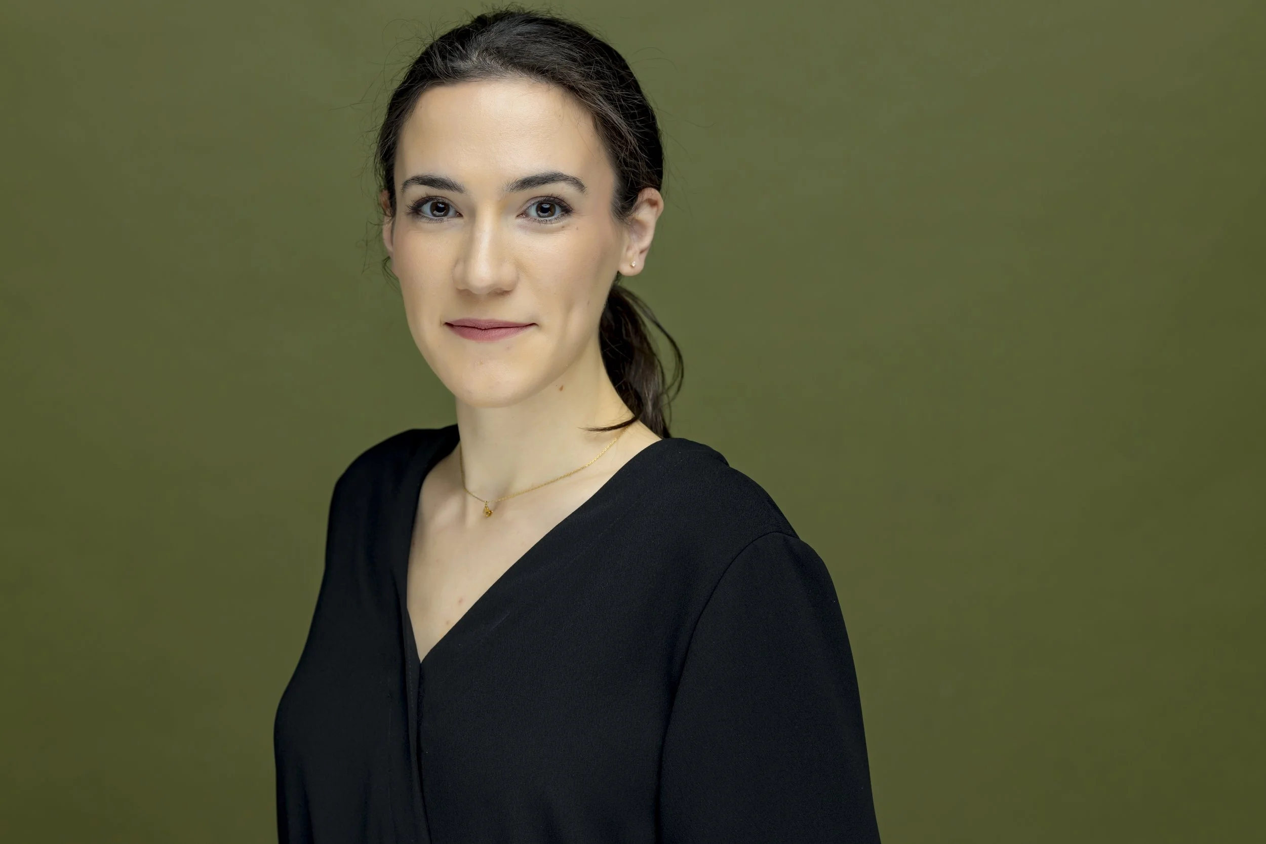 A woman with dark hair tied back, wearing a black top and a delicate gold necklace, standing against a plain green background.