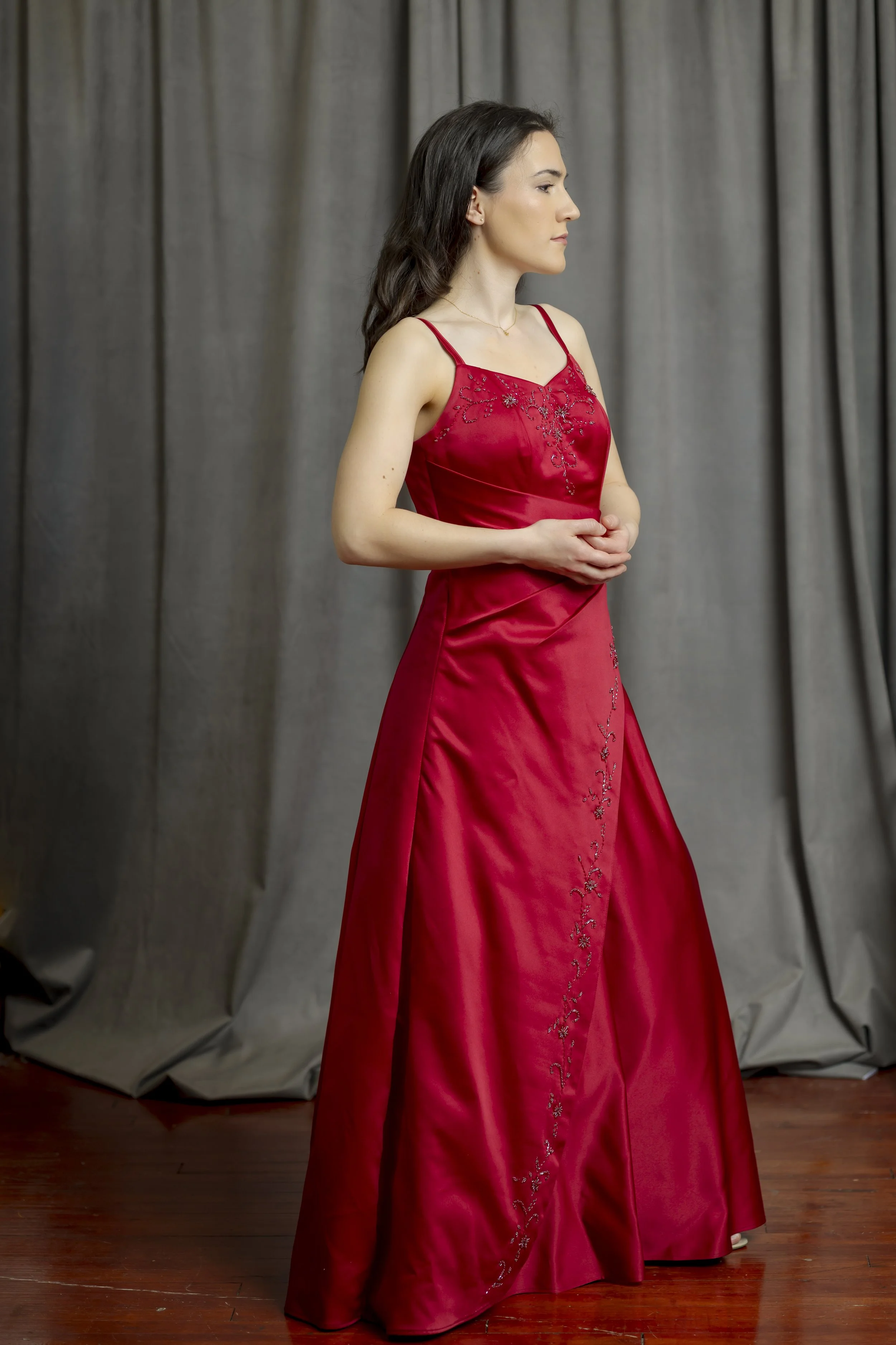 A woman in a red satin dress with floral embroidery around the neckline and down the front, standing with her hands clasped in front of her against a gray curtain background.
