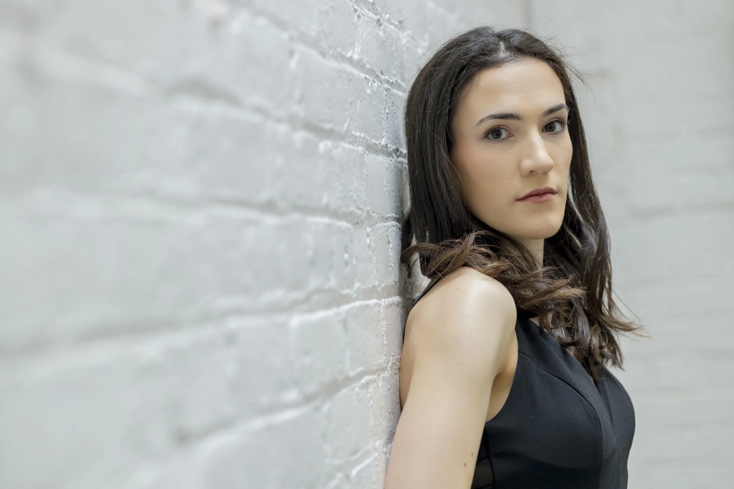 A woman with long dark brown hair and fair skin leaning against a white brick wall, looking at the camera with a neutral expression, wearing a sleeveless black top.