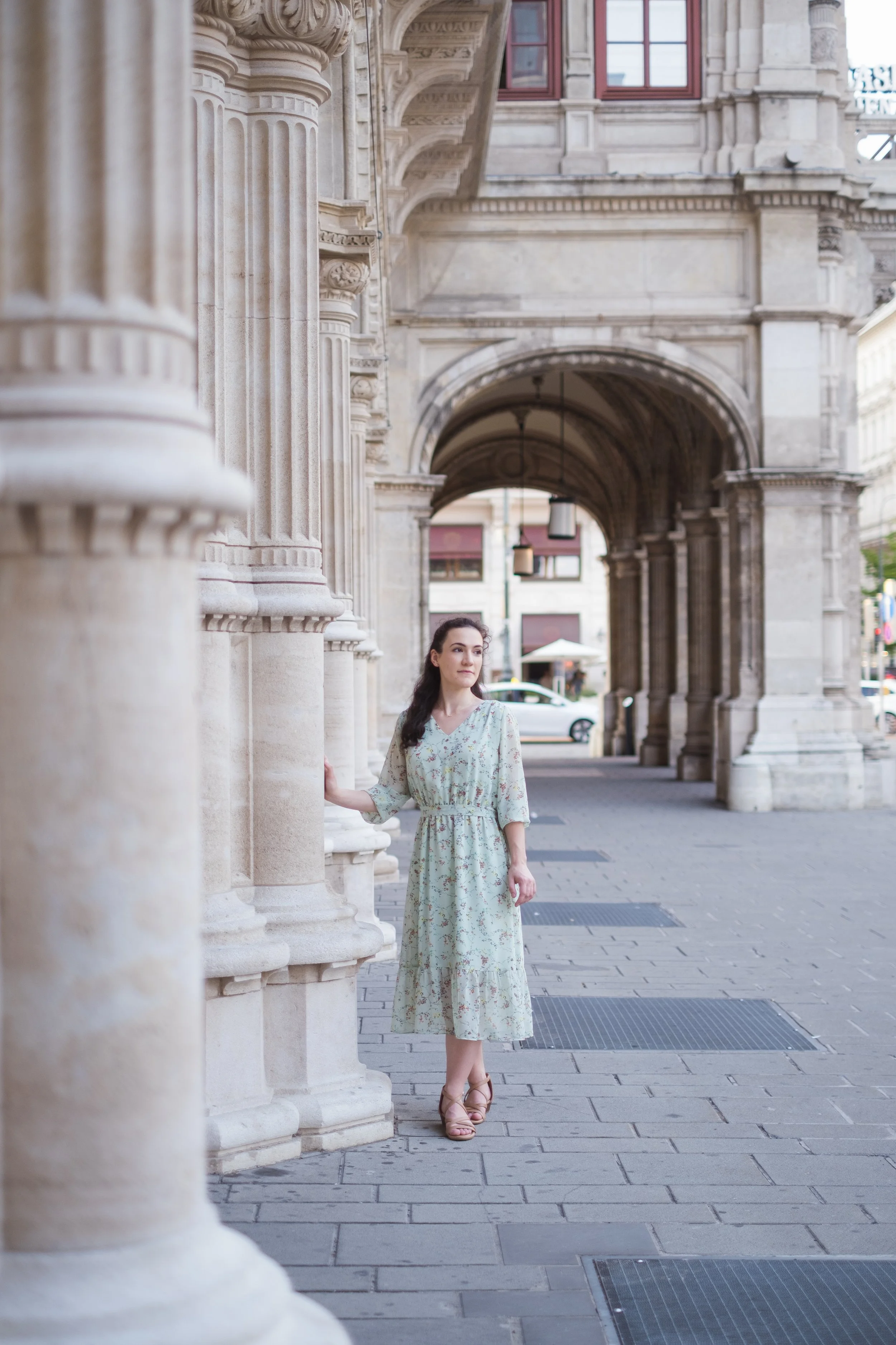 A woman in a light green floral dress and brown heels standing on a city sidewalk under historic stone arches.