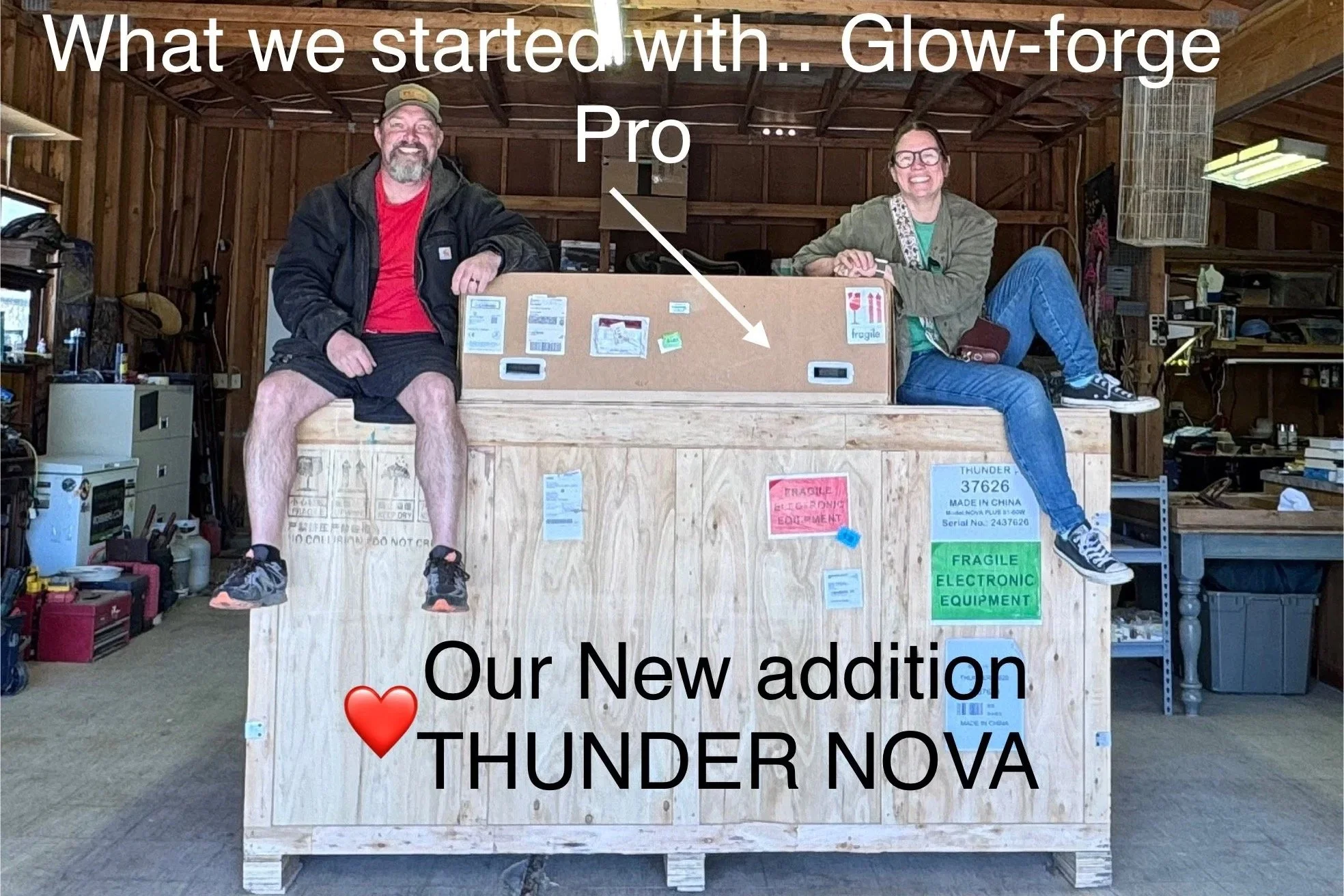 Two people sitting on a large wooden box in a workshop with various tools, equipment, and boxes around. The man on the left is wearing a cap, black jacket, red shirt, shorts, and sneakers. The woman on the right is wearing glasses, a green jacket, teal shirt, jeans, and sneakers. There is a large box labeled "THUNDER NOVA" with tags indicating fragile electronic equipment. The image has overlaid text that reads: "What we started with... Glow-forge Pro" at the top, and "Our New addition ❤️ THUNDER NOVA" at the bottom.