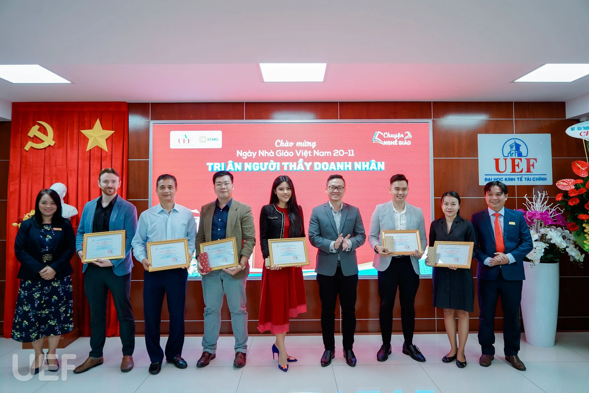 Group of nine people standing in a line, holding certificates, at a recognition event in front of a red digital display with Vietnamese text and logos, with a red curtain and a statue of a head in the background.