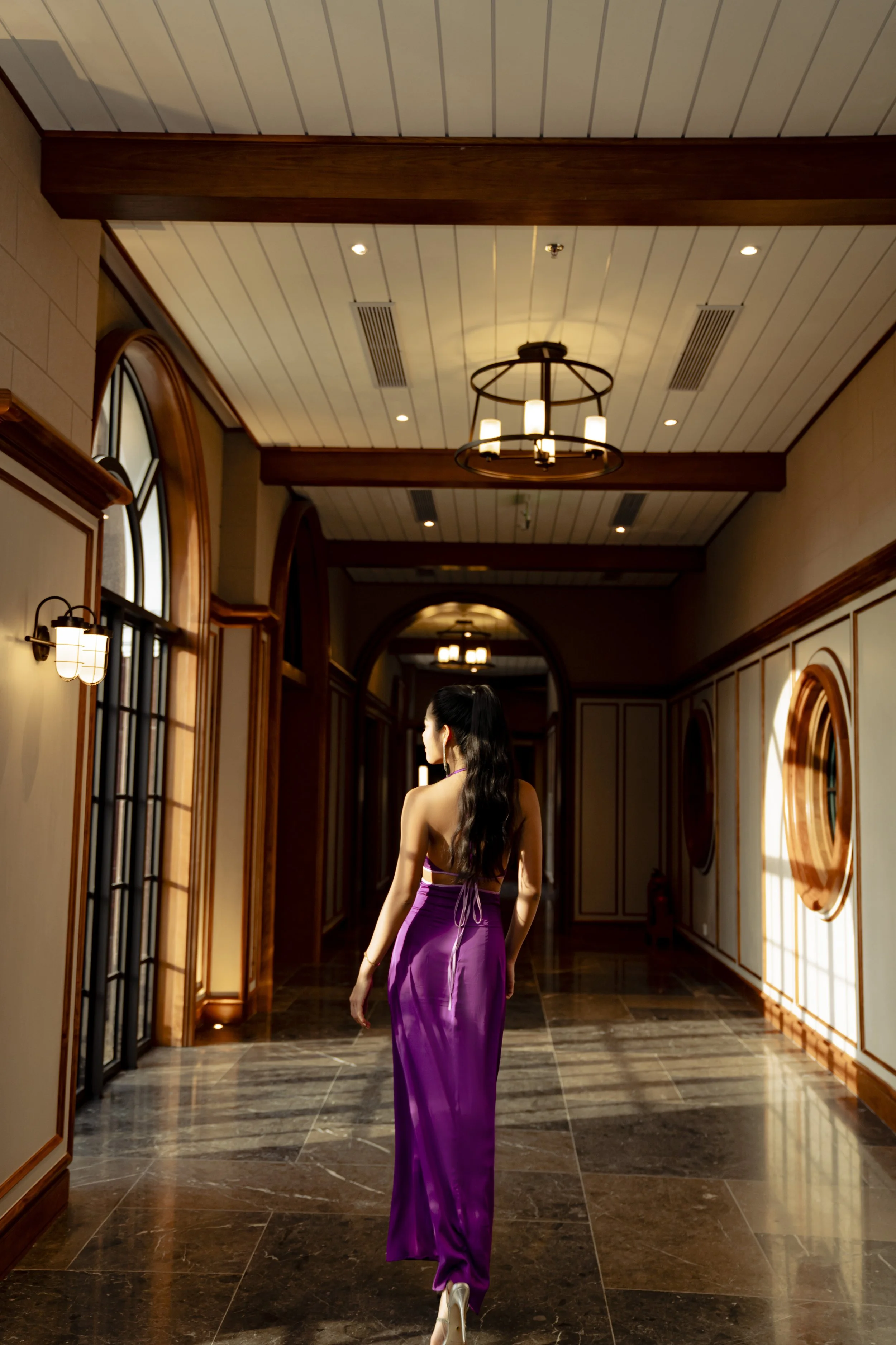 A woman in a purple dress walking down a marble-tiled hallway with chandeliers and wooden paneling.