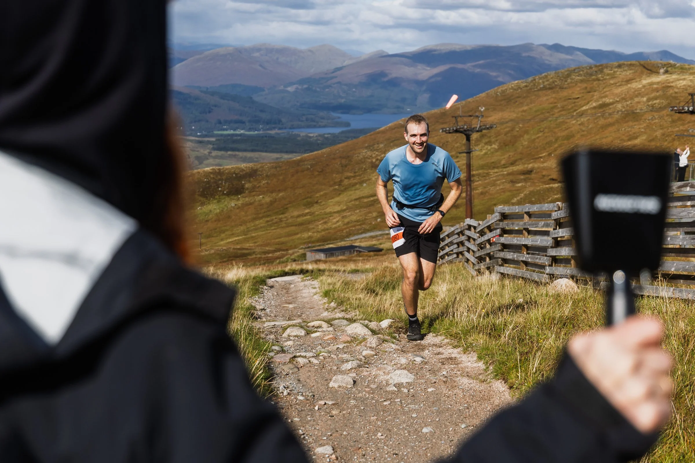 A man running on a rocky trail in a rural, mountainous landscape with hills, a lake, and a partly cloudy sky, as a person in the foreground holds a smartphone to record him.