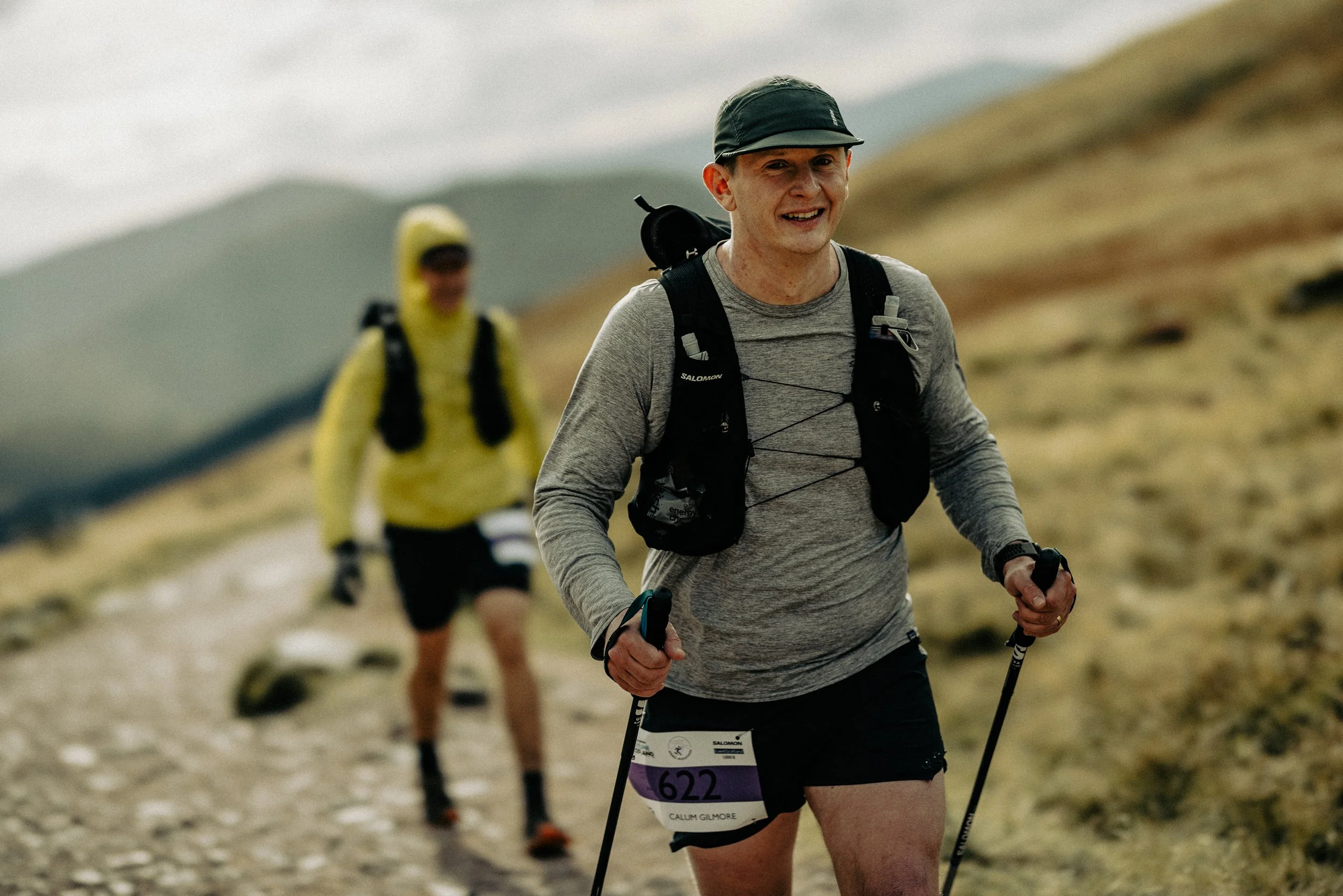 Two male hikers trail running on a mountain path with grassy terrain and distant mountains in the background. The man in the foreground is smiling, wearing a black hat, gray long-sleeve shirt, black shorts, and a black hydration vest, while the man i