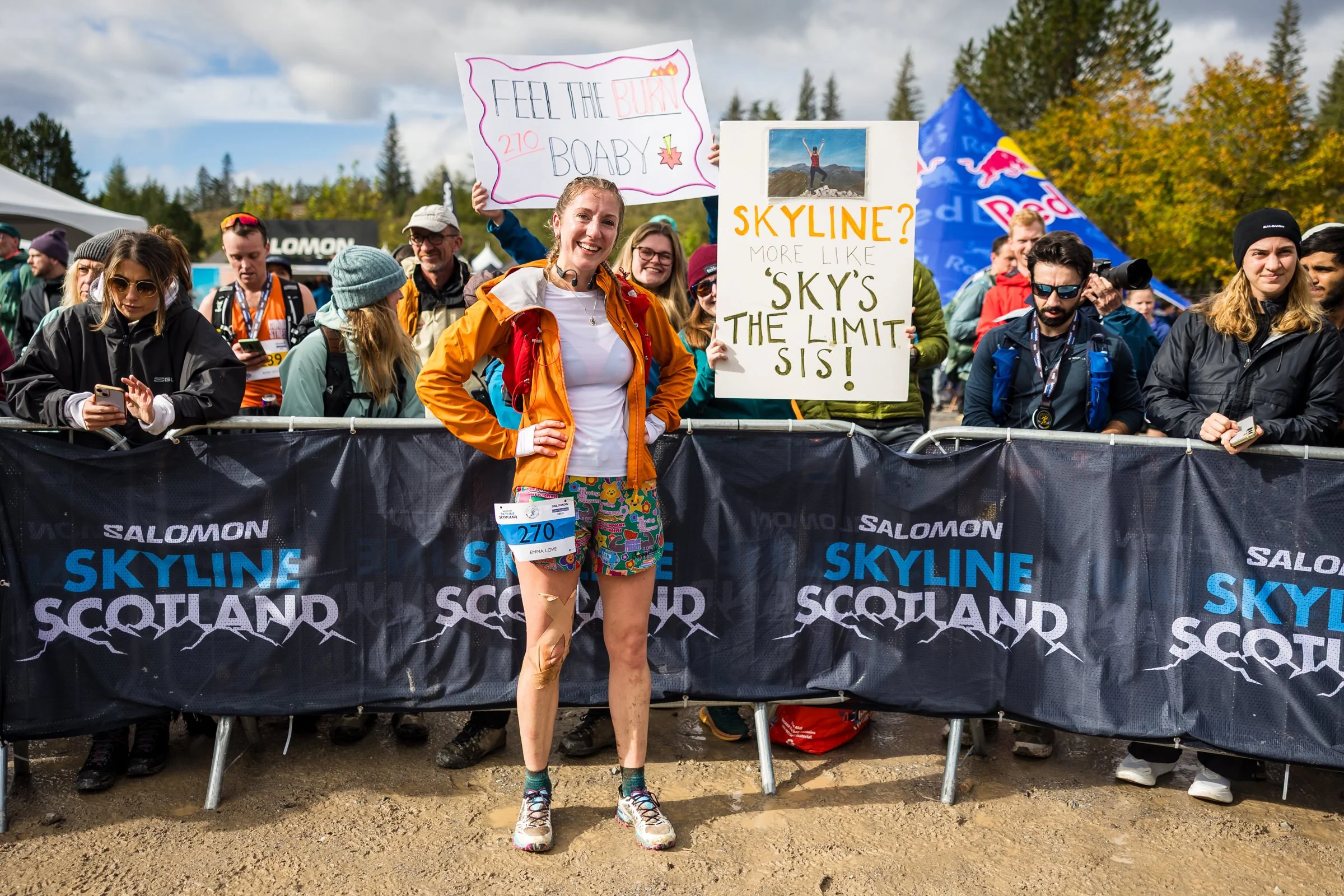 A woman smiling at a marathon event, with crowd and signs in the background, wearing a white shirt, orange jacket, colorful shorts, and sneakers.