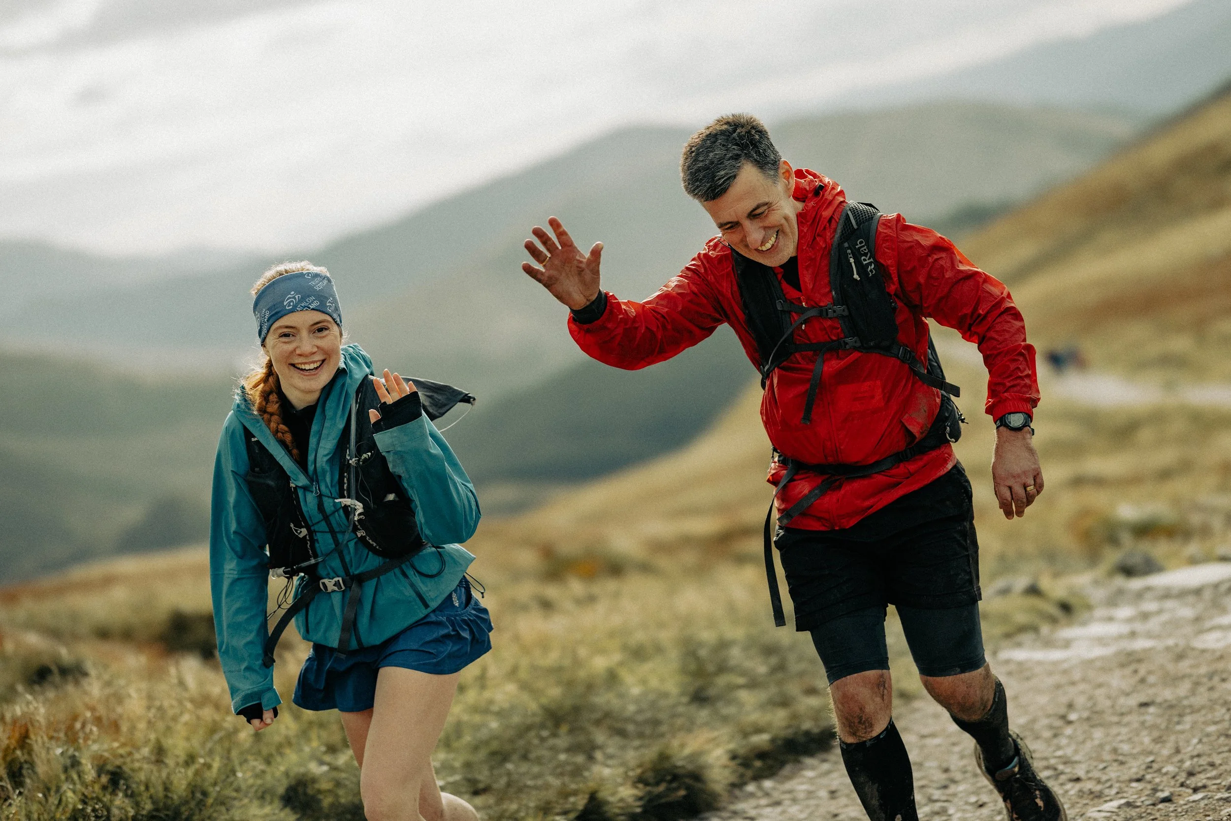 Two smiling hikers, a woman and a man, trail running on a mountain path with green hills in the background.