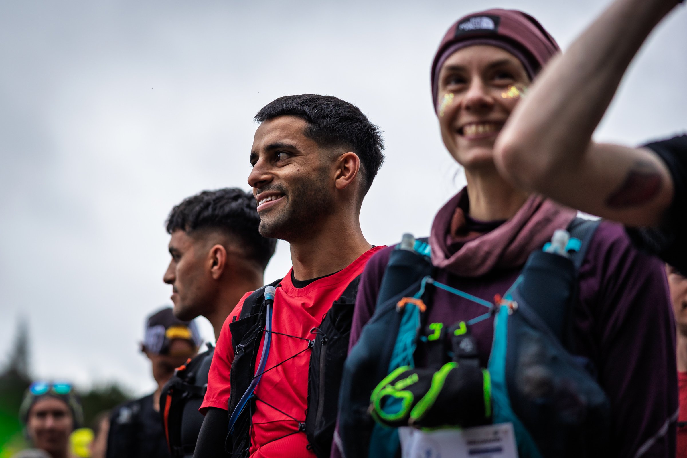 Group of smiling marathon runners standing outdoors before a race, wearing active gear and hydration packs.
