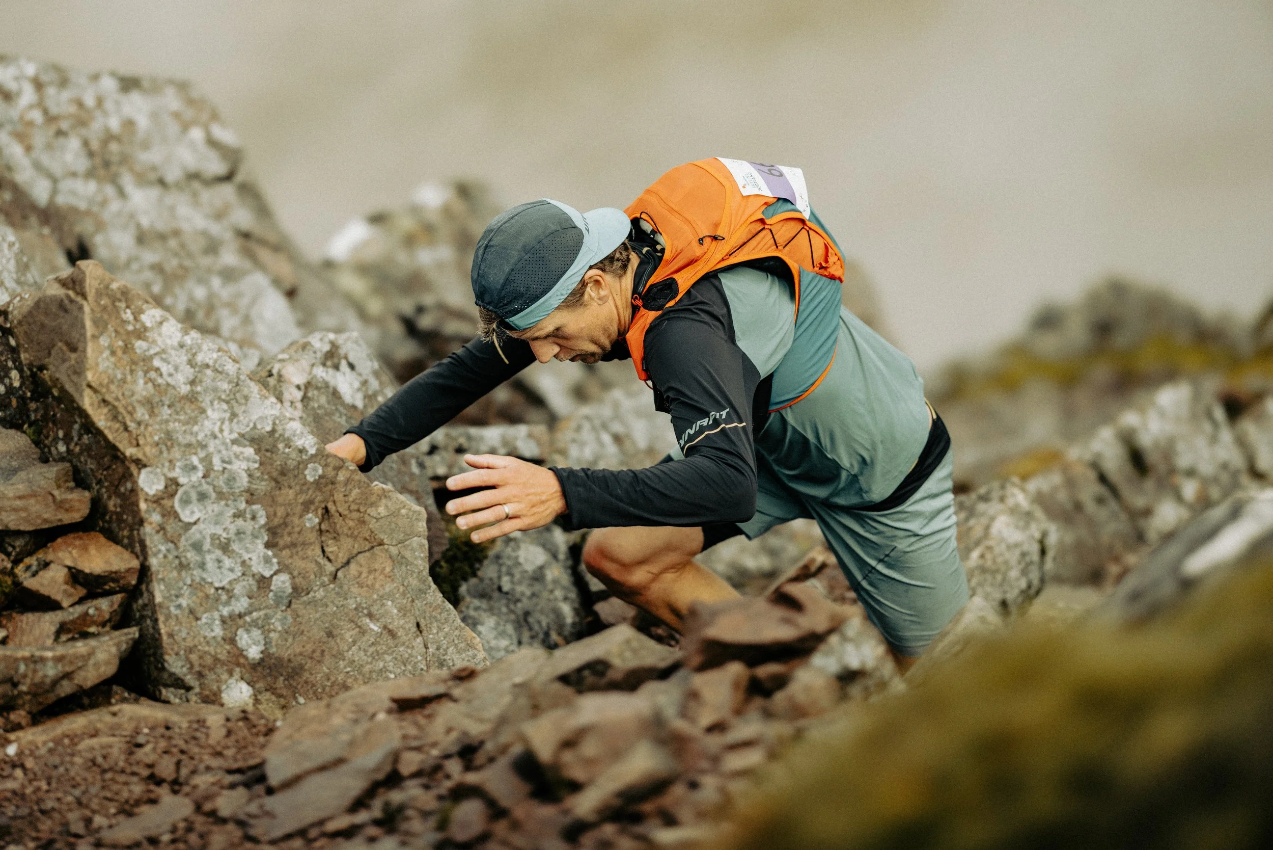 A male mountain climber ascending a rocky slope on a mountain trail, wearing outdoor gear and an orange backpack.