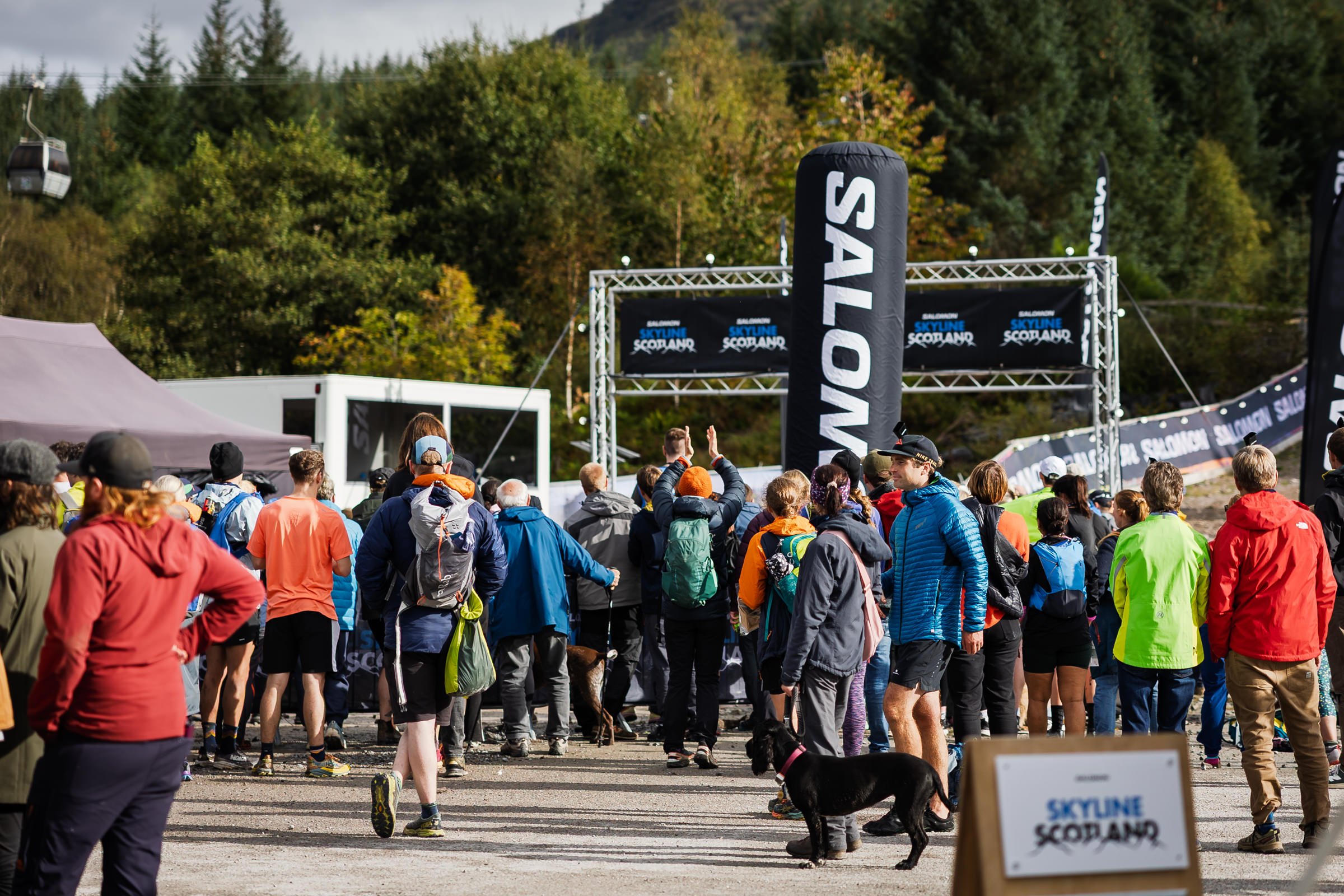 Crowd of people gathering outdoors at a marathon or race event in Scotland, with banners reading 'Skyline Scotland' and large SALOMON flags, set against a backdrop of trees and hills.