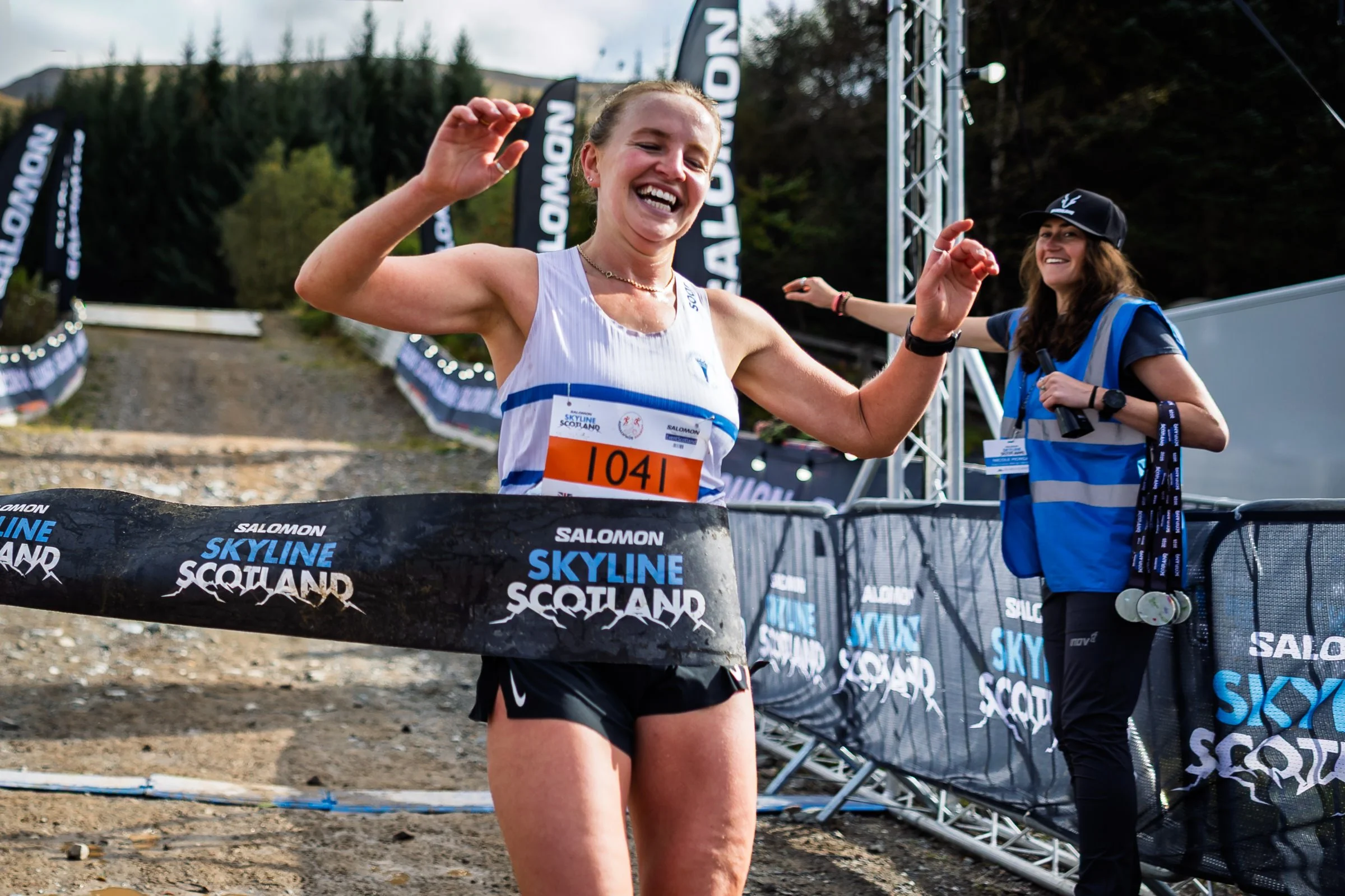 A female runner is crossing the finish line, smiling and raising her arms in celebration, with a race bib numbered 104. A woman in a blue vest and cap stands behind her, holding a microphone and wearing a lanyard and watch. The background features banners and signs with the words "Salomon Skyline Scotland" and a mountainous outdoor setting.