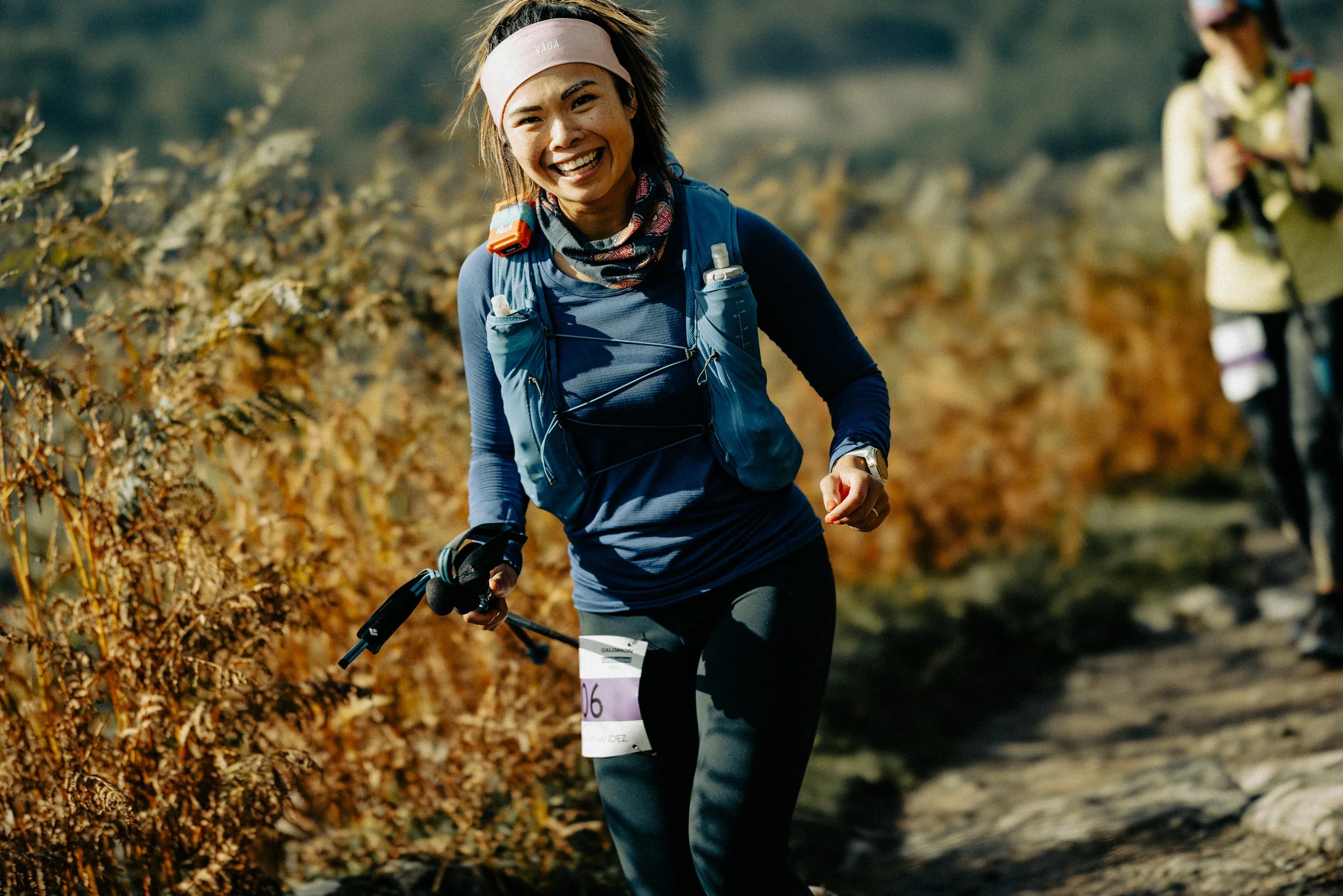 A woman running on a trail through a forest with autumn-colored foliage, wearing a headband, a blue long-sleeve shirt, black pants, and carrying a backpack and trekking poles.