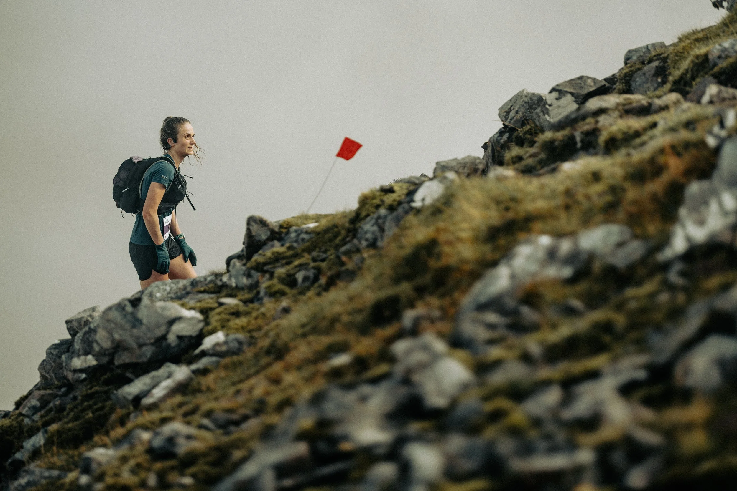 A woman dressed in hiking gear with a backpack, walking up a rocky, moss-covered mountain slope during cloudy weather, with a small red flag in the background.
