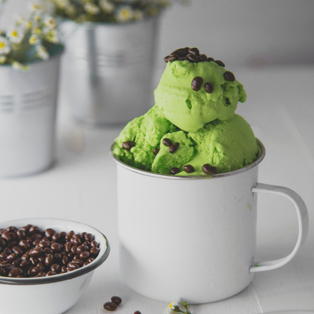 Scoops of bright green ice cream topped with coffee beans in a white mug on a white surface, with a bowl of coffee beans in the foreground and metal containers with flowers in the background.