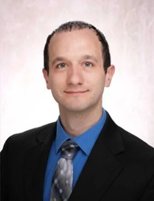A man with short dark hair, wearing a light blue dress shirt and matching tie, smiling outdoors with blurred trees and sunlight in the background.