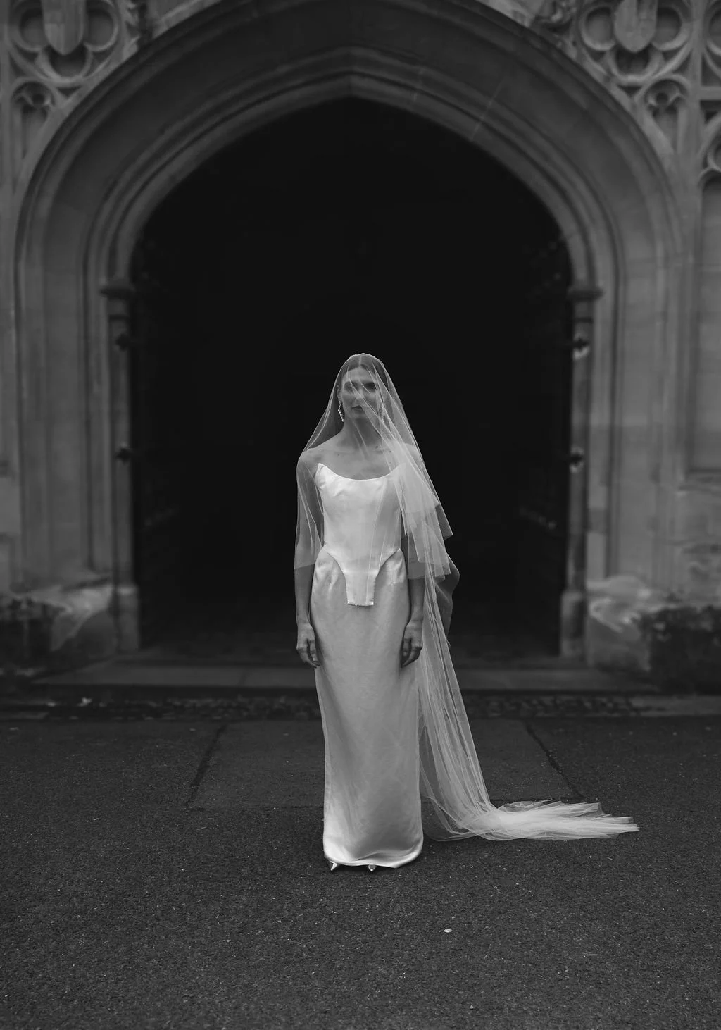 A woman in a wedding dress and veil stands in front of a dark arched entrance in black and white.
