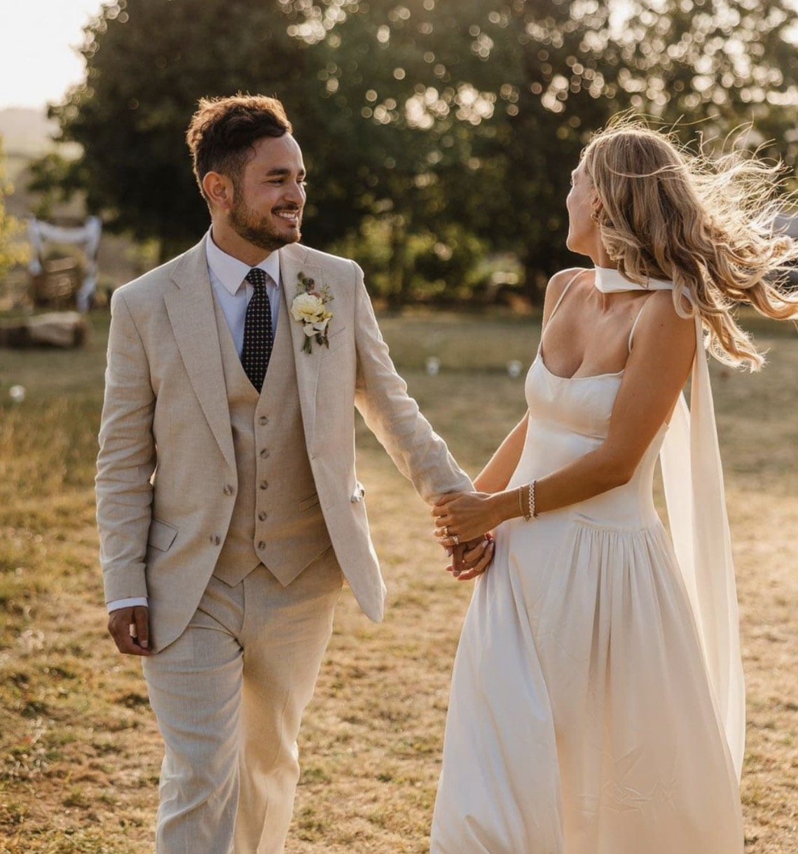 A wedding couple holding hands outdoors during sunset, the groom wearing a beige suit with a flower boutonniere and the bride in a white dress with flowing hair.