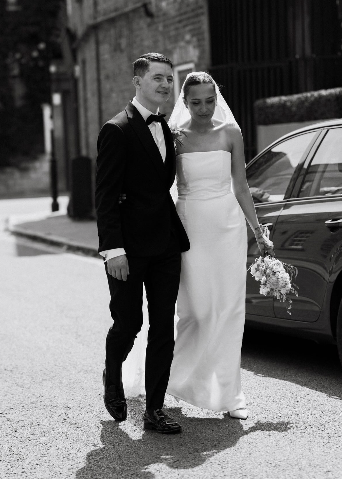 A black-and-white photo of a bride and groom walking together on a street. The groom is wearing a tuxedo and the bride is in a strapless wedding dress holding a bouquet of flowers.
