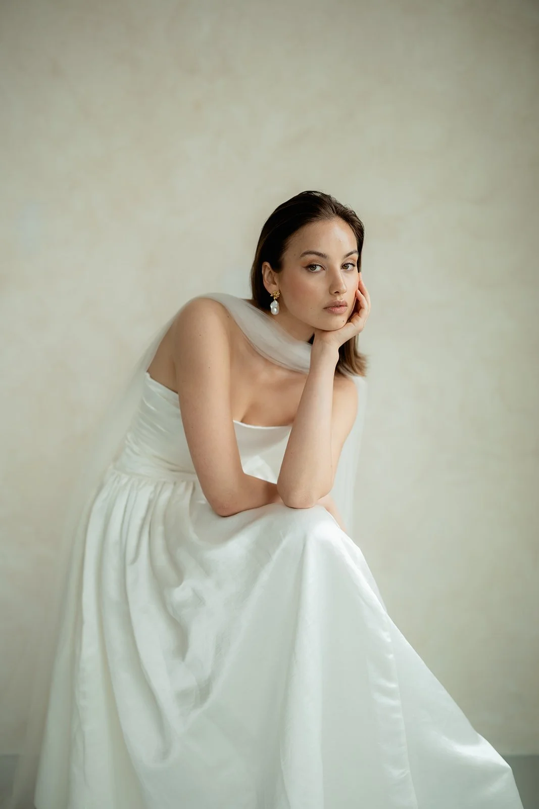 A woman in a white dress with a sheer neckline, sitting against a plain light background, resting her chin on her hand and looking at the camera.