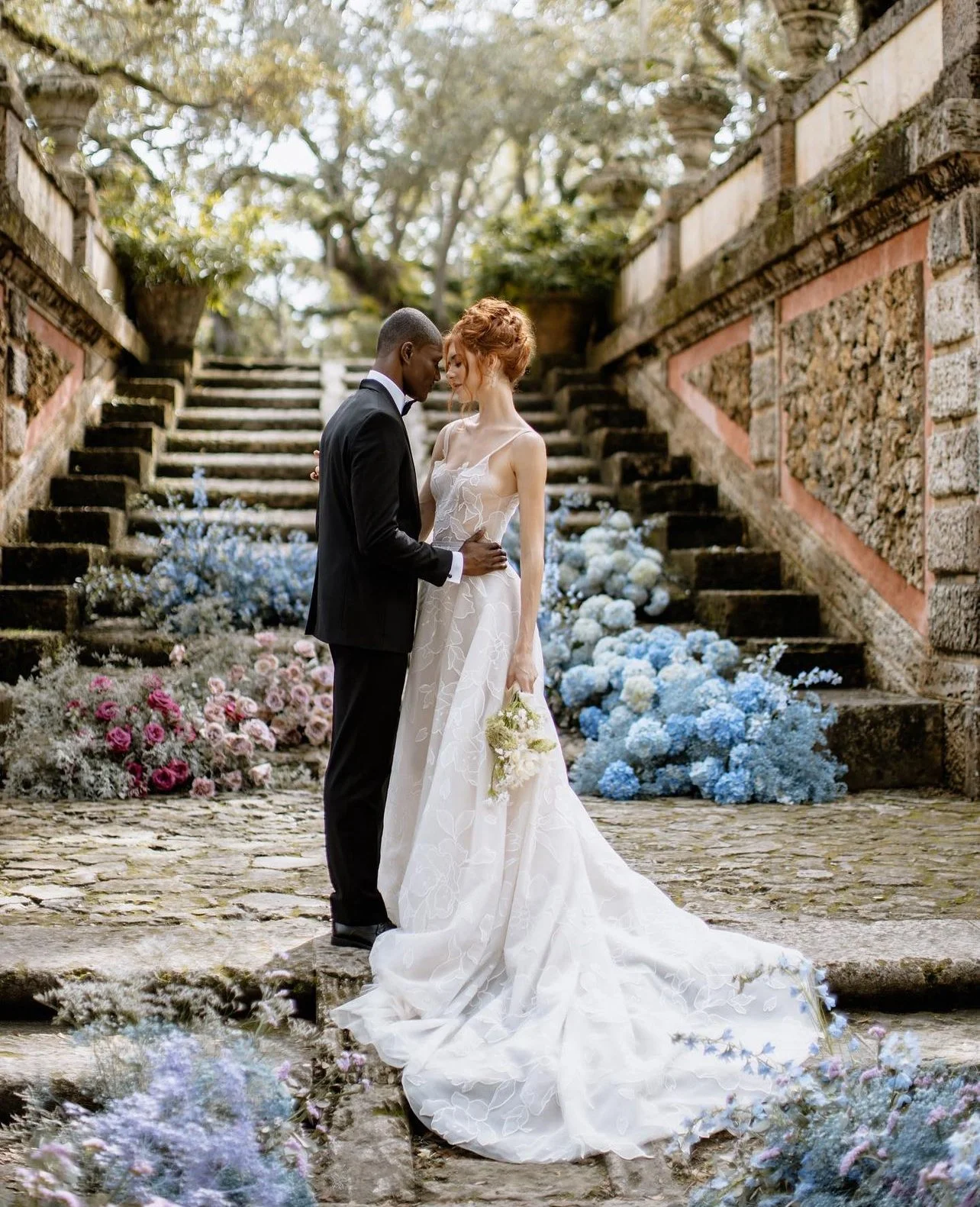 Un couple en robe de mariage et costume noir se tient dans un cadre rustique avec des escaliers en pierre, entouré de fleurs colorées et d'arbres au fond.