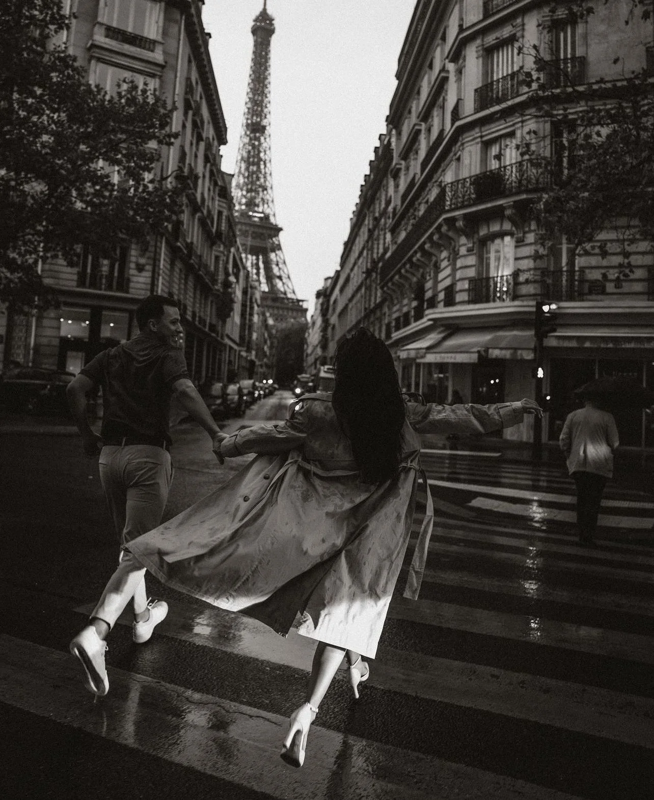 Une scène urbaine à Paris avec la Tour Eiffel en arrière-plan. Deux personnes, un homme et une femme, dansent ou jouent dans la rue détrempée sous la pluie, en noir et blanc.
