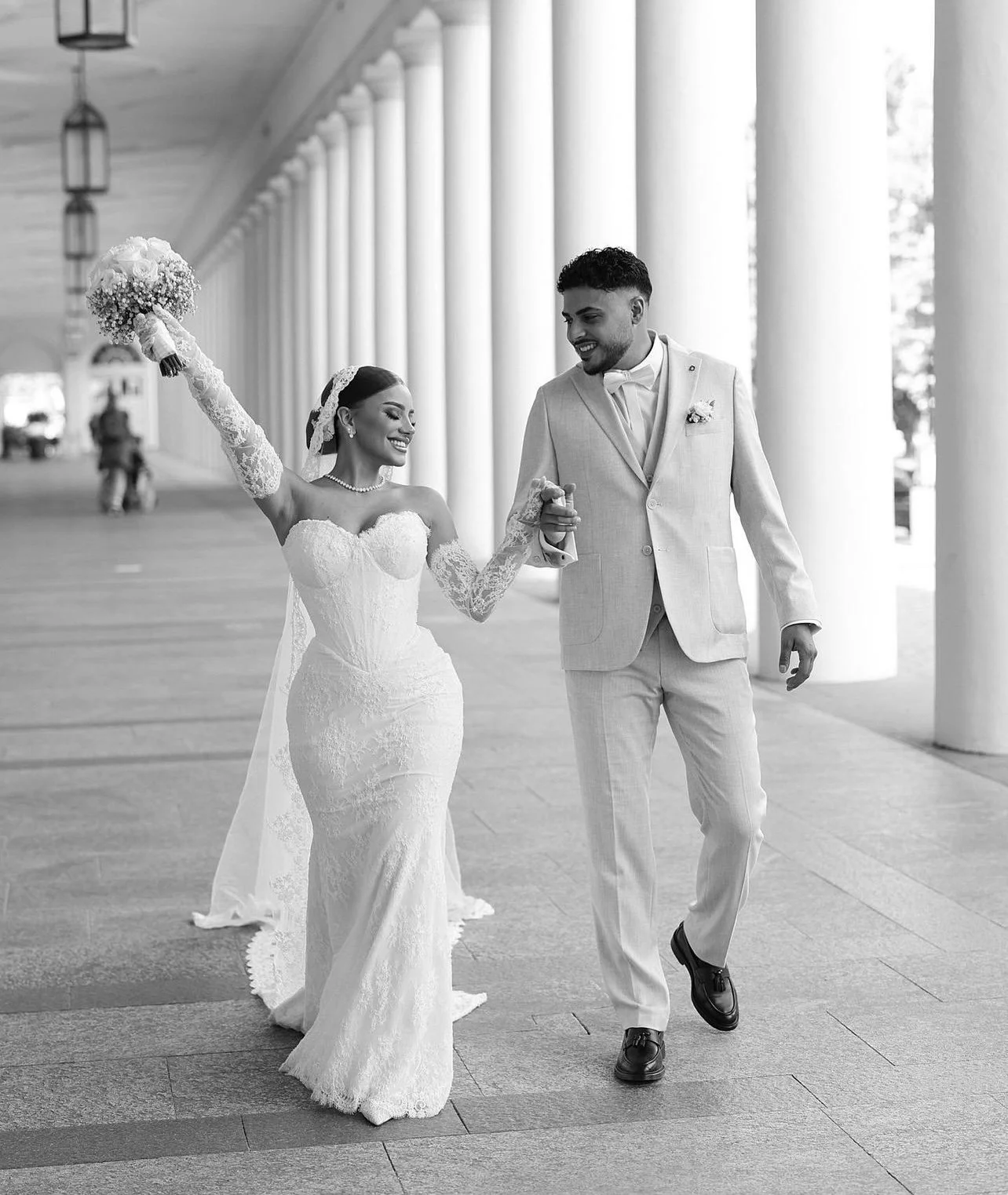 Mariage élégant avec une mariée en robe de mariée et un marié en costume, marchant sous une arcade avec des colonnes.