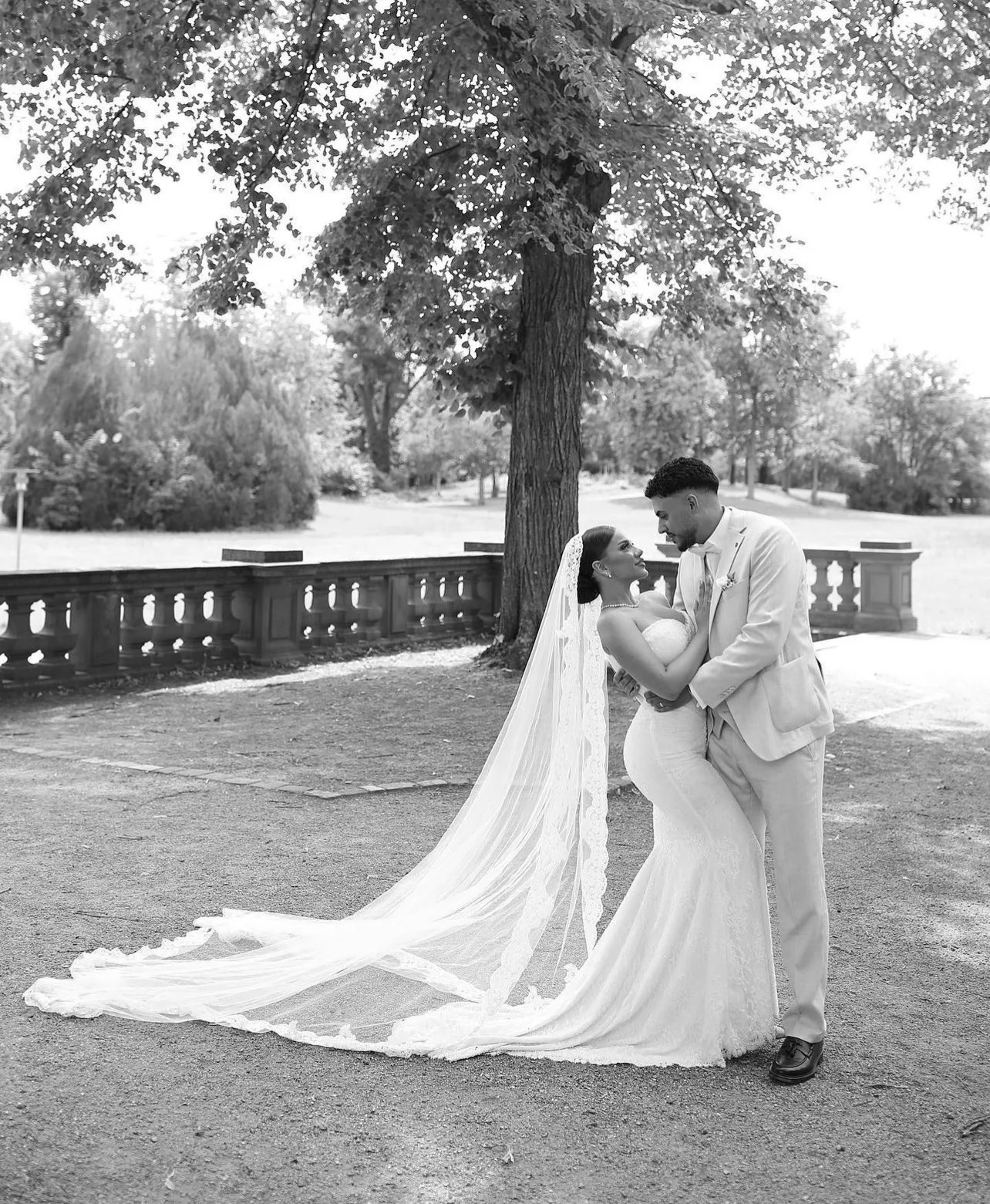 Un couple de mariés dans un parc, en noir et blanc, avec la mariée portant une robe de mariage longue et un voile, et le marié en costume clair, face à face, sous un grand arbre.