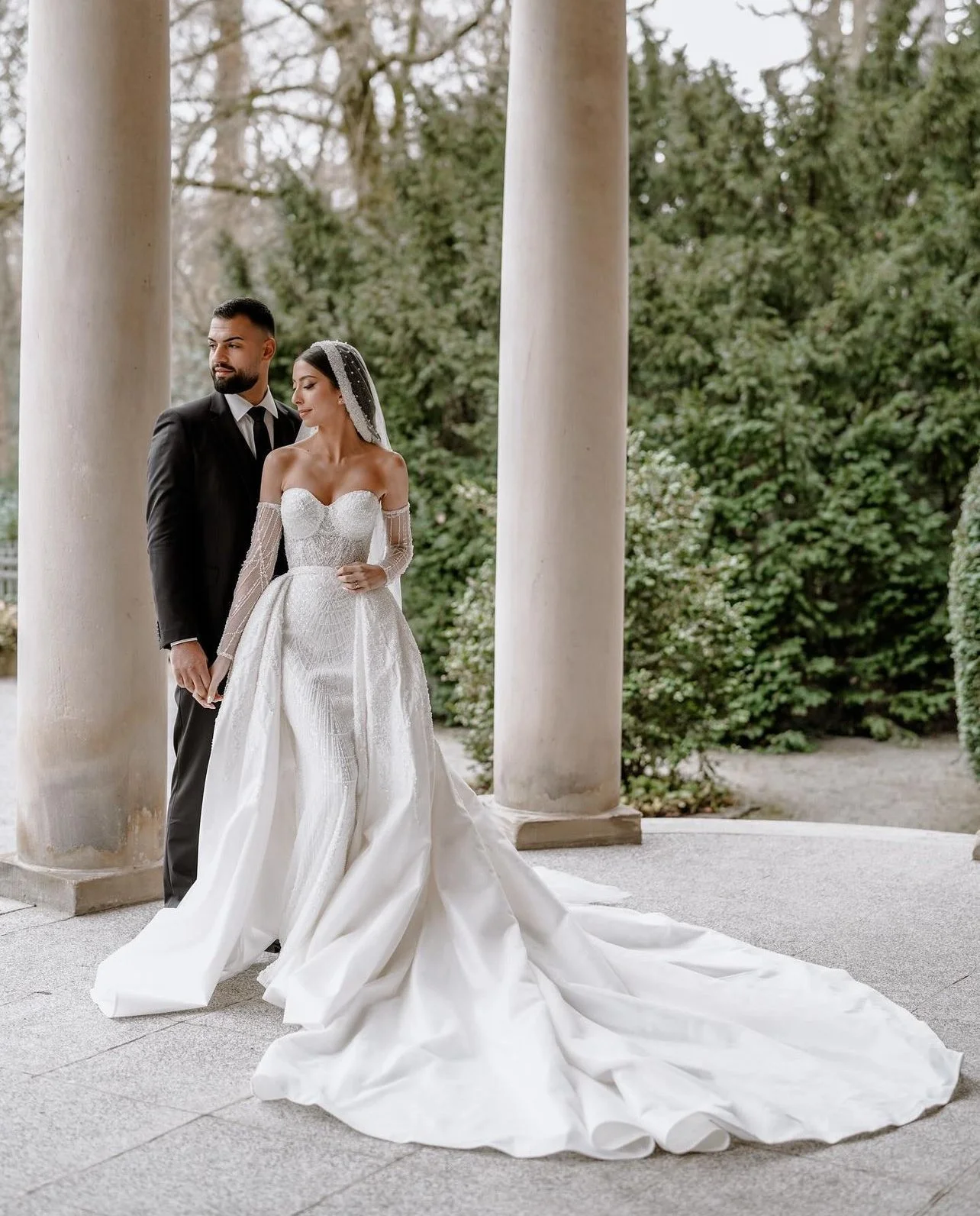 Un couple en robe de mariage et costume, posant devant des colonnes en pierre et des arbres verts, lors d'une séance photo nuptiale.