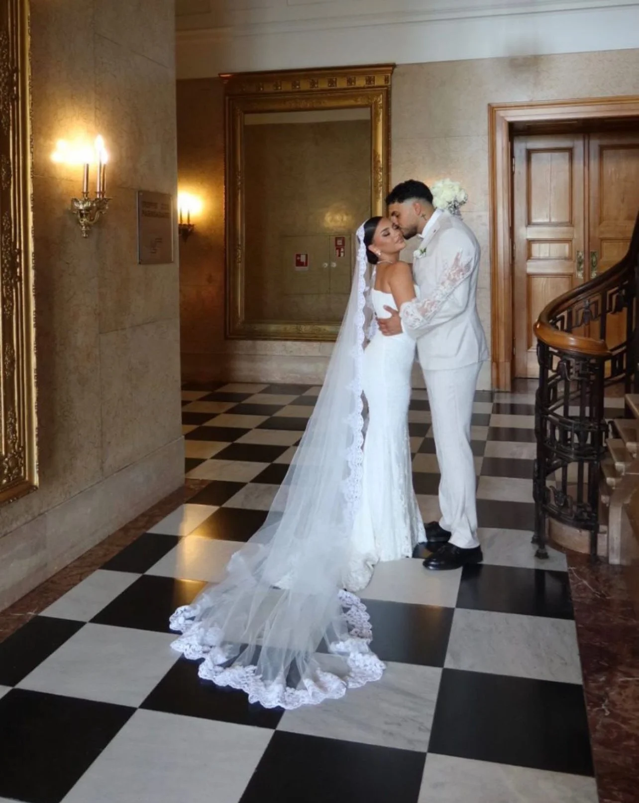 Un couple de mariés en tenue de mariage s'embrassant dans une salle élégante avec sol en carreaux de marbre noir et blanc, murs en pierre et un grand miroir doré.