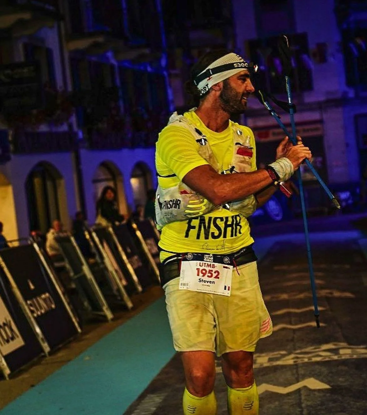 A male marathon runner, wearing yellow athletic clothing and a headlamp, is crossing the finish line at night, holding trekking poles, with a bib number 1952.