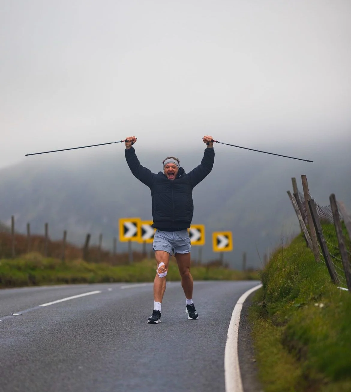 A man celebrating with arms raised while running on a curving road in a rural area, holding two hiking poles, wearing a jacket, shorts, and a bandana, with a cloudy sky in the background.