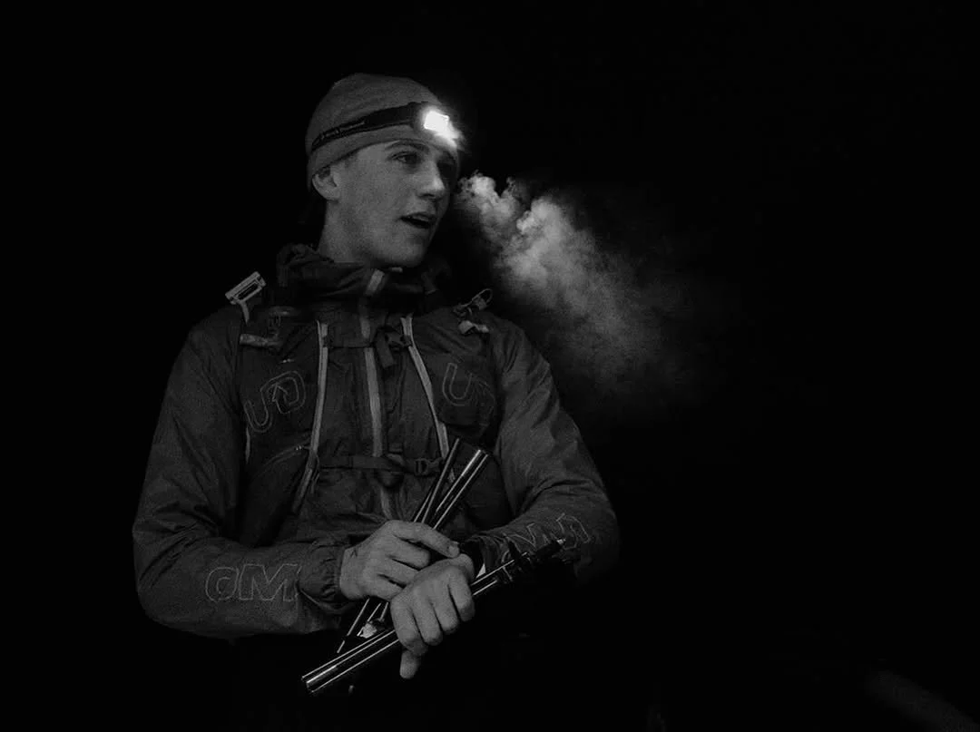 Black and white photo of a young woman wearing a headlamp and outdoor gear, inhaling or exhaling vapor or breath in cold weather.
