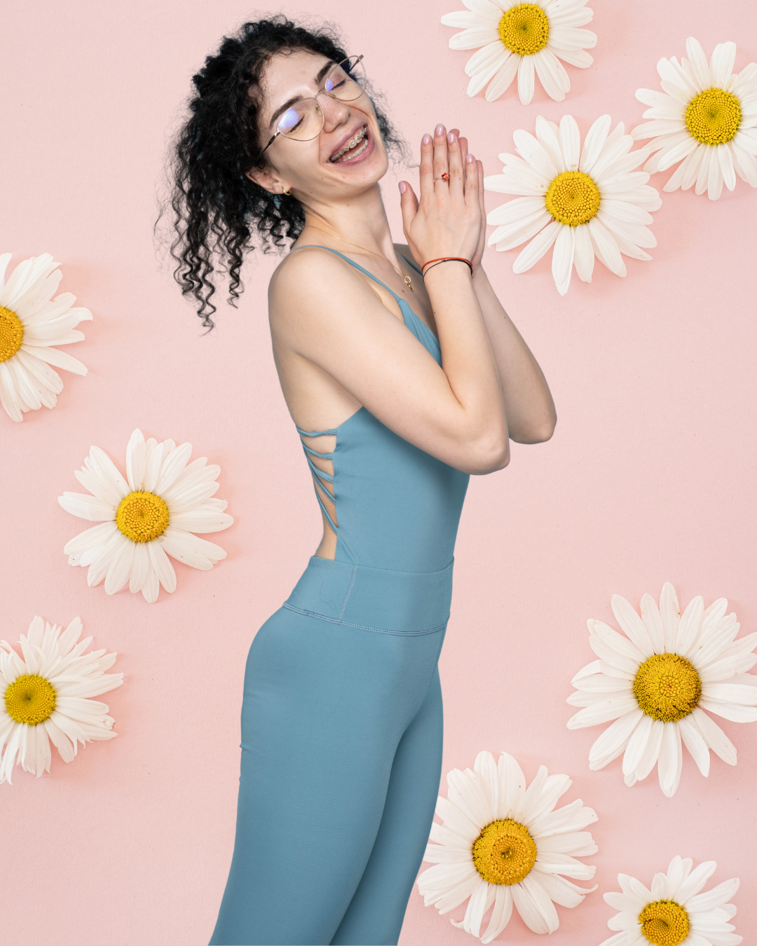 A young woman with curly black hair, glasses, and braces, wearing a blue athletic outfit, standing against a pink background decorated with white daisies. She is smiling with her eyes closed and hands together in a prayer position.