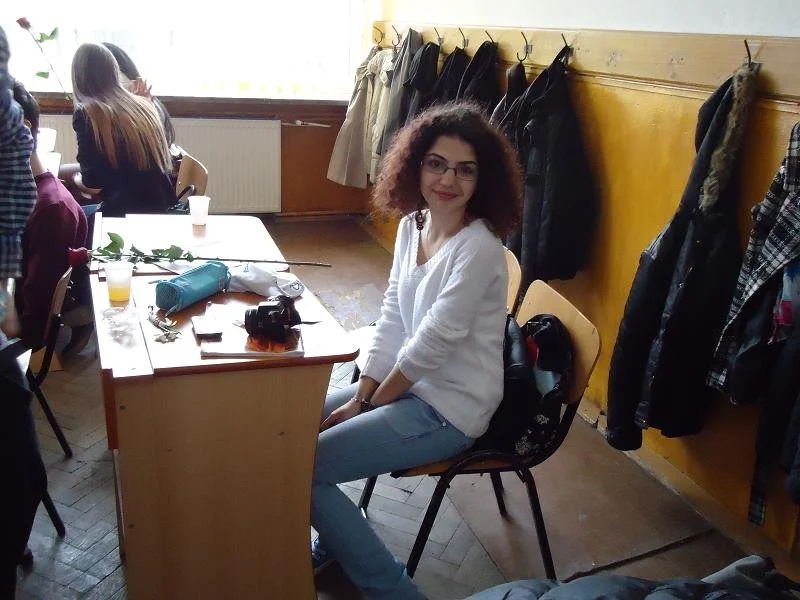 A young woman with curly hair and glasses sitting on a chair at a table in a room with coat hooks on the wall, smiling at the camera. Several coats hang on the hooks, and other people are seated at the table in the background.