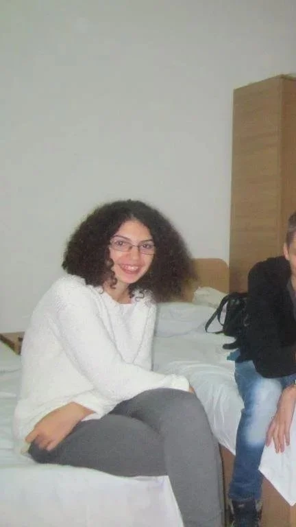 A woman with curly dark hair, wearing glasses and a white sweater, sitting on a bed with a smile.