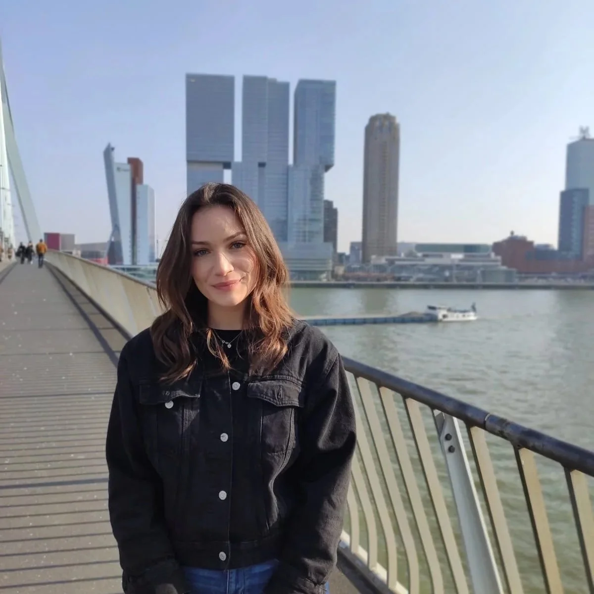 A young woman with shoulder-length brown hair and a black denim jacket standing on a bridge over a river in a city with modern skyscrapers in the background.